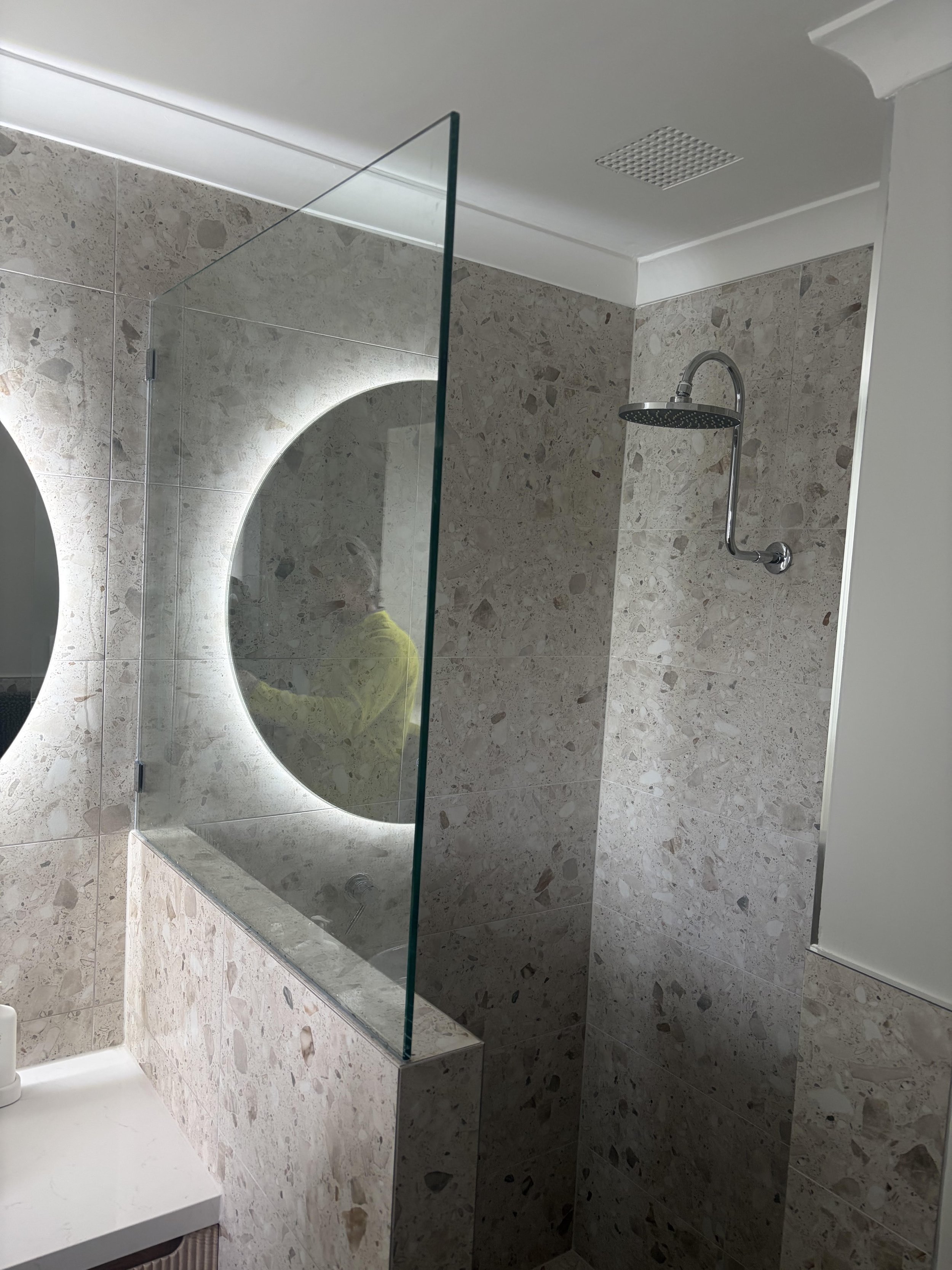 Modern bathroom with a shower that has a black shower head, a mirror with backlighting, beige and brown tile walls, a white ceiling with a vent, and a portion of a white countertop with a striped towel.