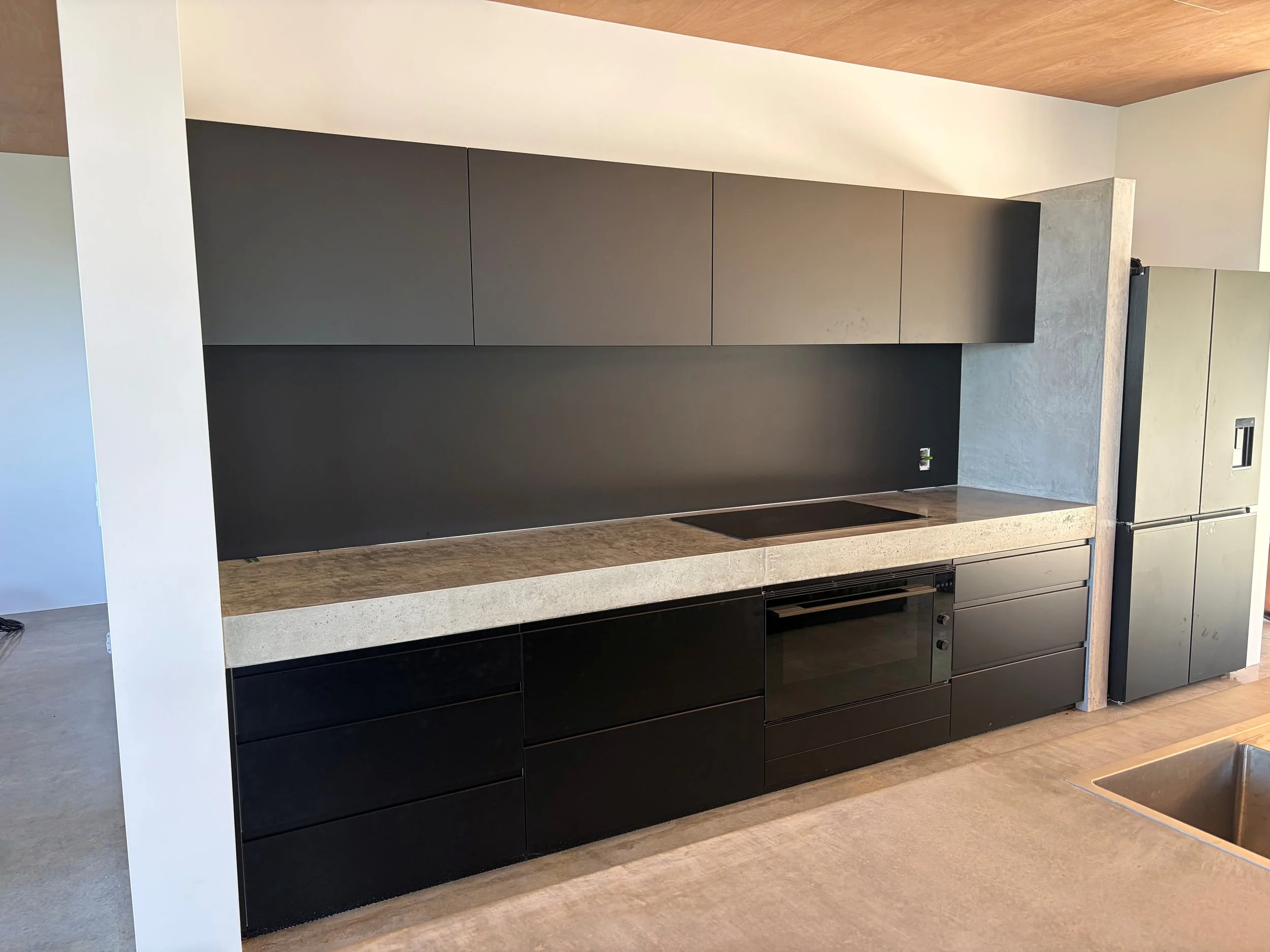 Modern black and gray kitchen cabinetry with a white and concrete countertop, located in a living space with wooden flooring and a light-colored wall.