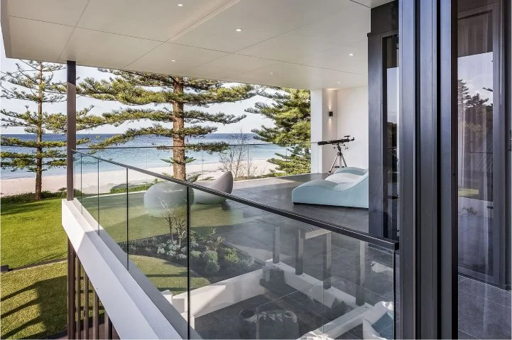Modern balcony overlooking a grassy area and water with trees, featuring a white lounge chair, a telescope, and a glass railing.