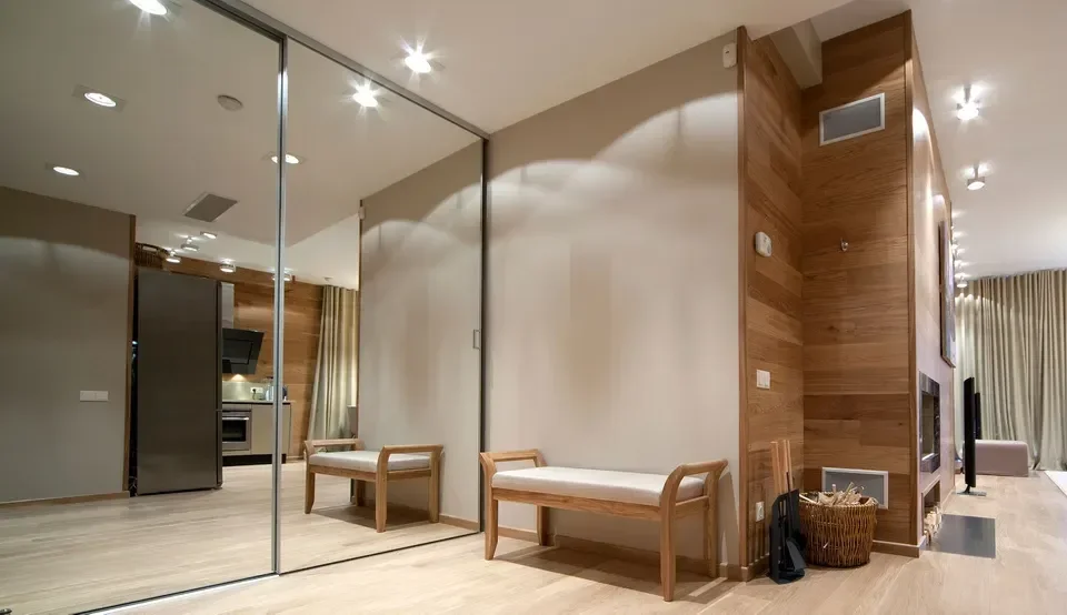Modern living space with wooden paneled walls, two wooden benches with white cushions, a glass sliding door, and a glimpse of the kitchen area in the background.