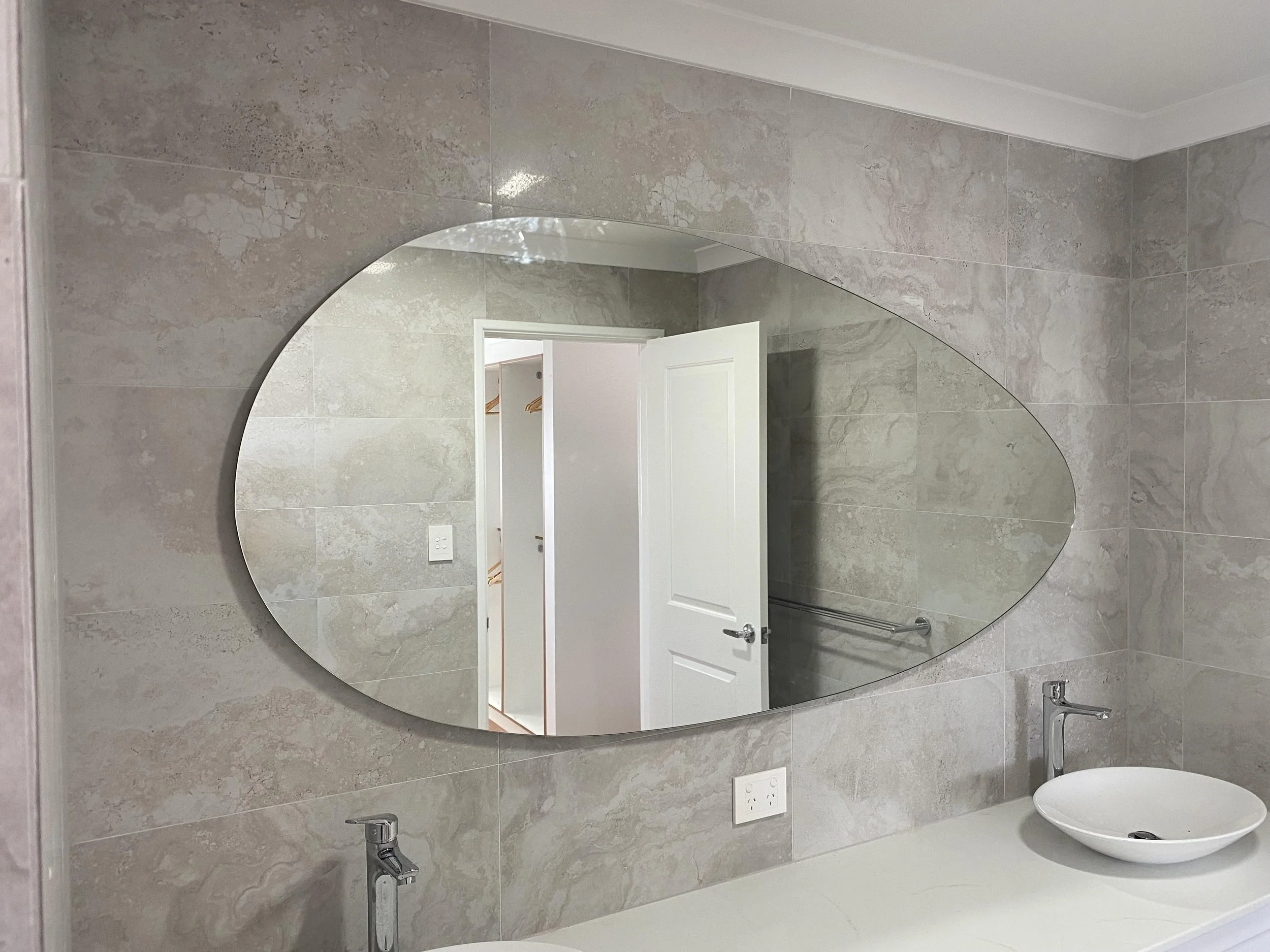 Bathroom with grey marble tiles on the wall, large oval mirror, white door, and a white vessel sink with a modern faucet.