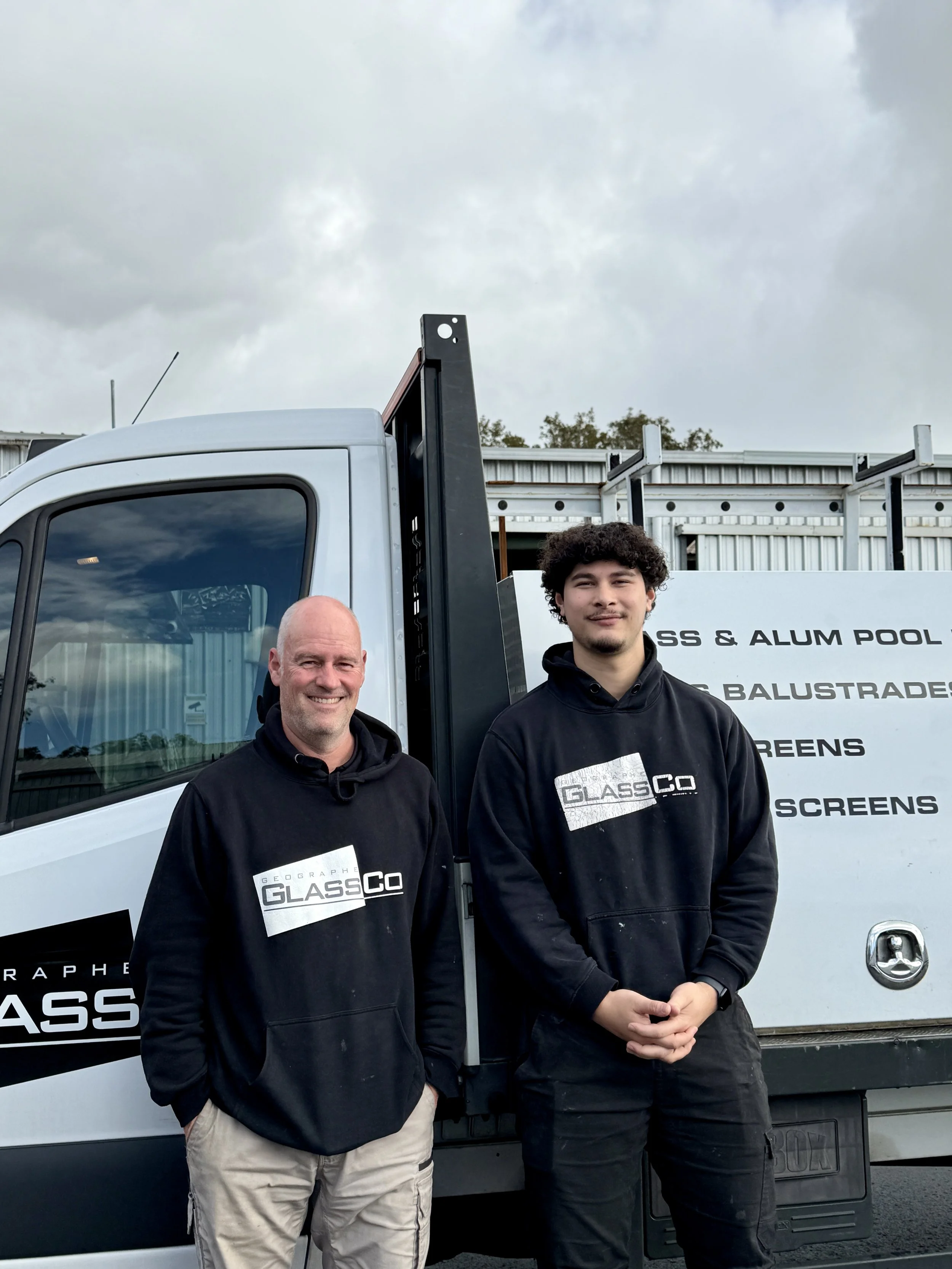 Two men standing in front of a white truck with signage for a glass and aluminum company. The truck's side panel has text related to glass, aluminum, and balustrades. The older man on the left has a bald head, a smile, and is wearing a black hoodie with the logo 'GEOGRAPHIC GLASS CO.' The younger man on the right has curly hair, a slight smile, and is wearing a black hoodie with the logo 'GLASS CO.' They are outdoors on a cloudy day with a fence and some trees in the background.