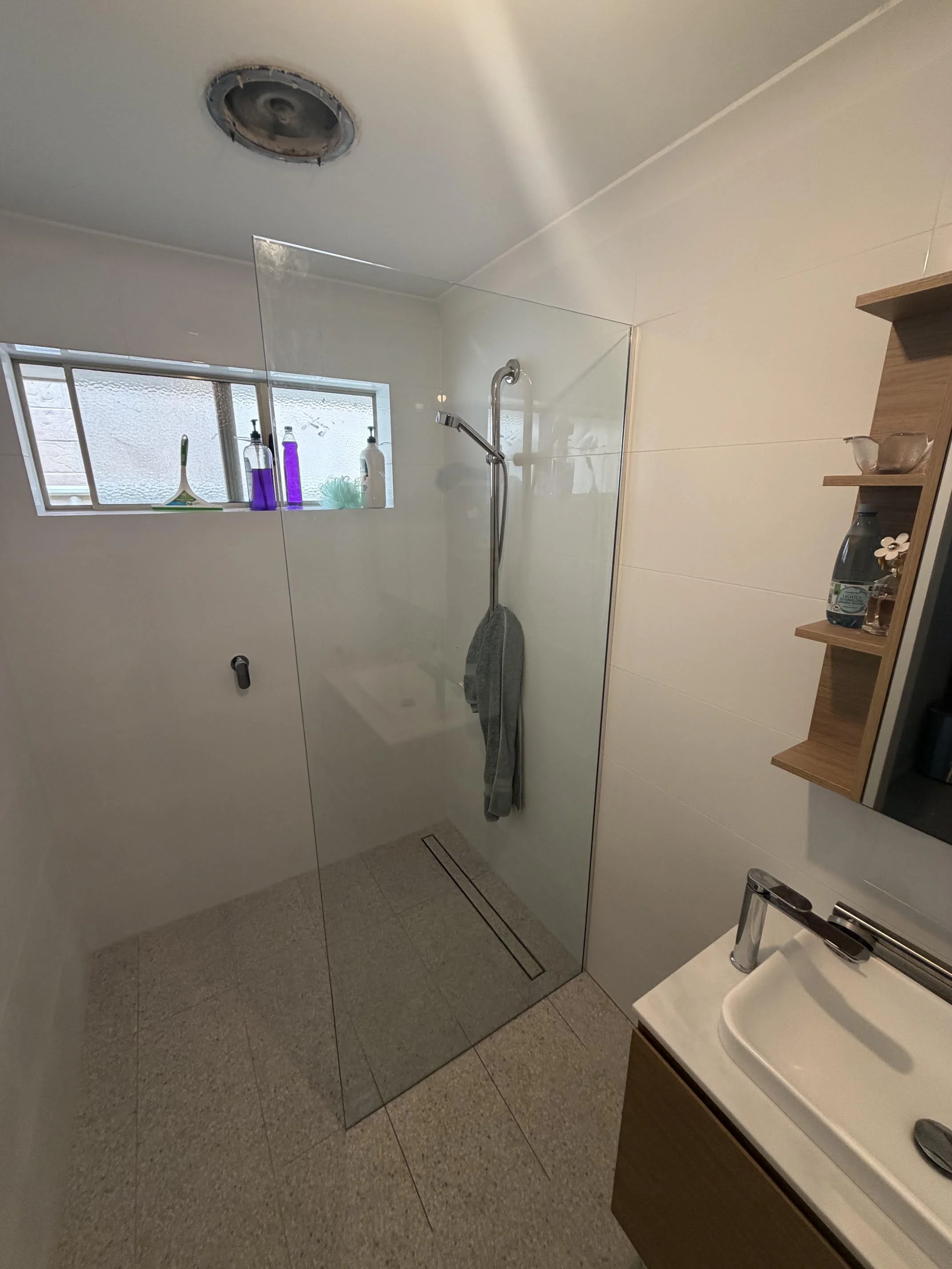 Modern bathroom with a glass shower enclosure, small window with bottles and a soap dispenser, a wall-mounted shelf, and a sink with a wooden cabinet and a mirror.