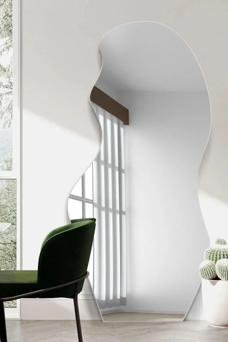 Decorative mirror shaped like a profile of a face on a white wall, with a green chair and a window with sheer curtains in a modern interior.