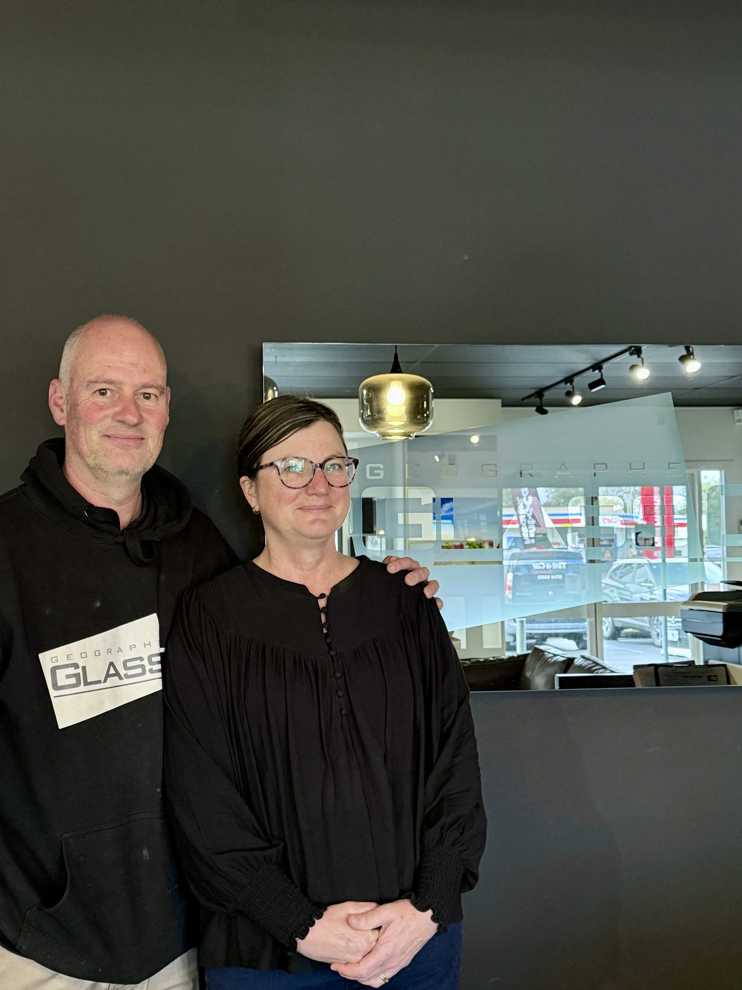 Two people, a man and a woman, standing indoors. The man has a bald head and is wearing a black jacket with a 'Geograph Glass' tag. The woman has short dark hair, glasses, and is wearing a black top. They are standing close together in front of a window with a frosted glass sign, and there is a ceiling light fixture above them.