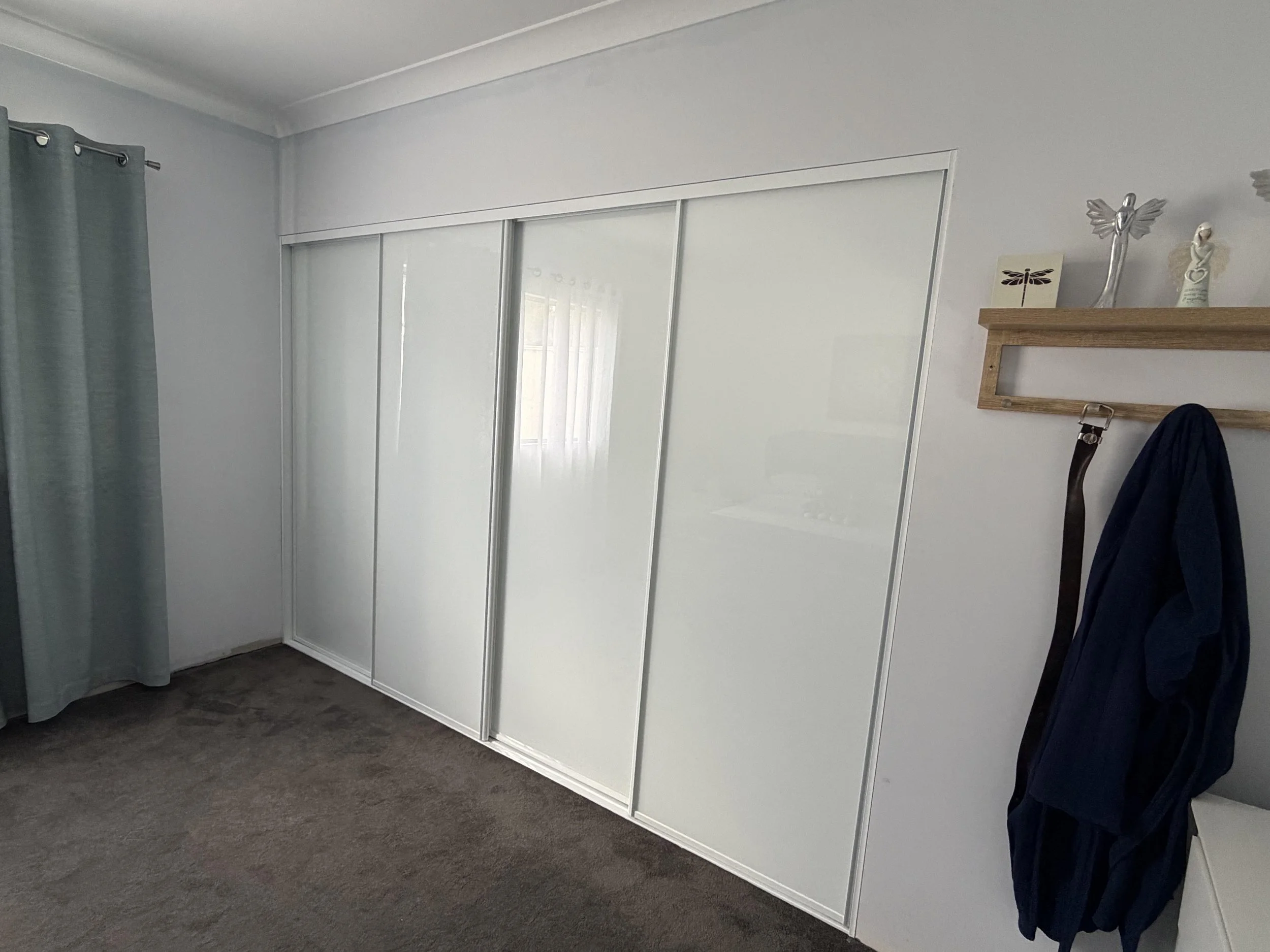A bedroom with a large white sliding door closet, light blue curtains on the left, gray carpeted floor, and a wooden coat rack on the right with some decorative items and a belt hanging from it. A dark jacket is hanging on the coat rack.