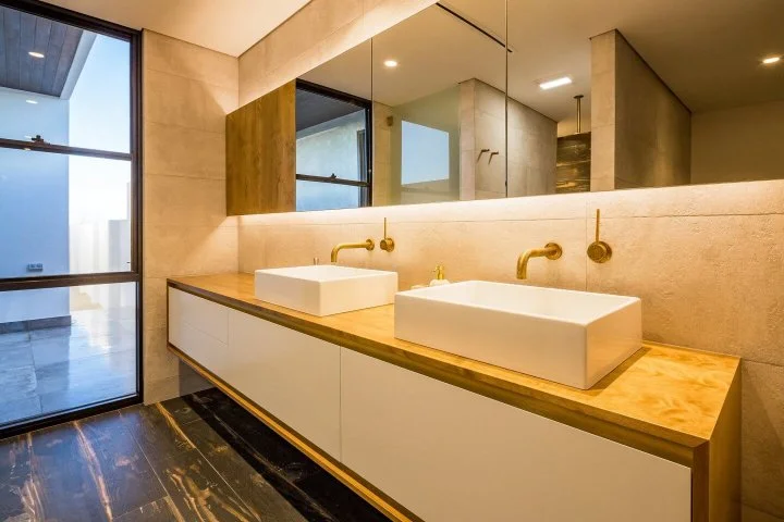 Modern bathroom with a double vanity featuring two rectangular vessel sinks, gold wall-mounted faucets, a large mirror, and a window letting in natural light.