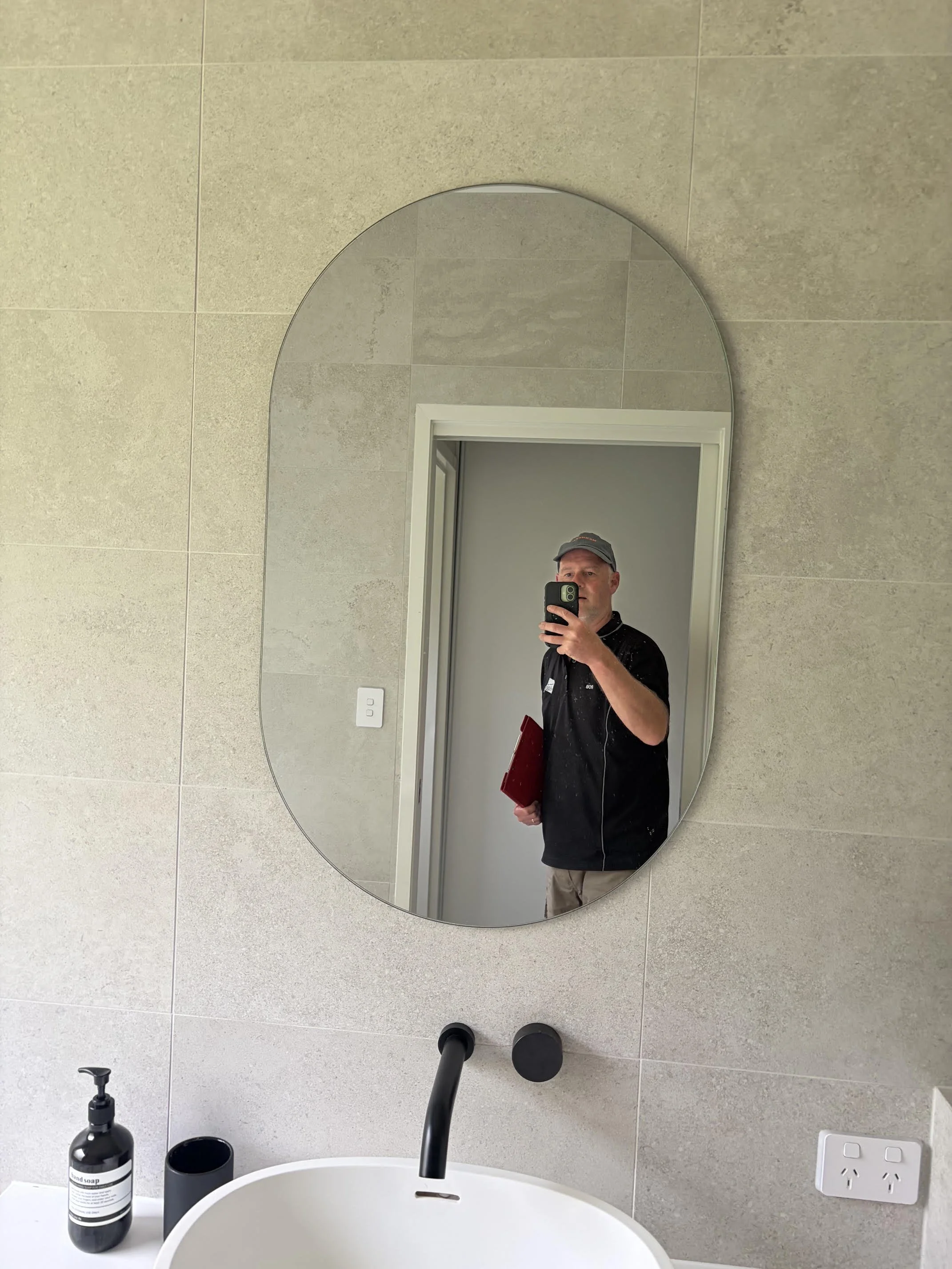 A bathroom mirror reflects a man holding a phone to take a selfie. The bathroom has beige tiled walls, a black soap dispenser, and a white sink.