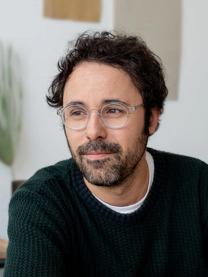 A man with dark curly hair and glasses looking thoughtful, sitting indoors with a blurred background that includes a potted plant and artwork.