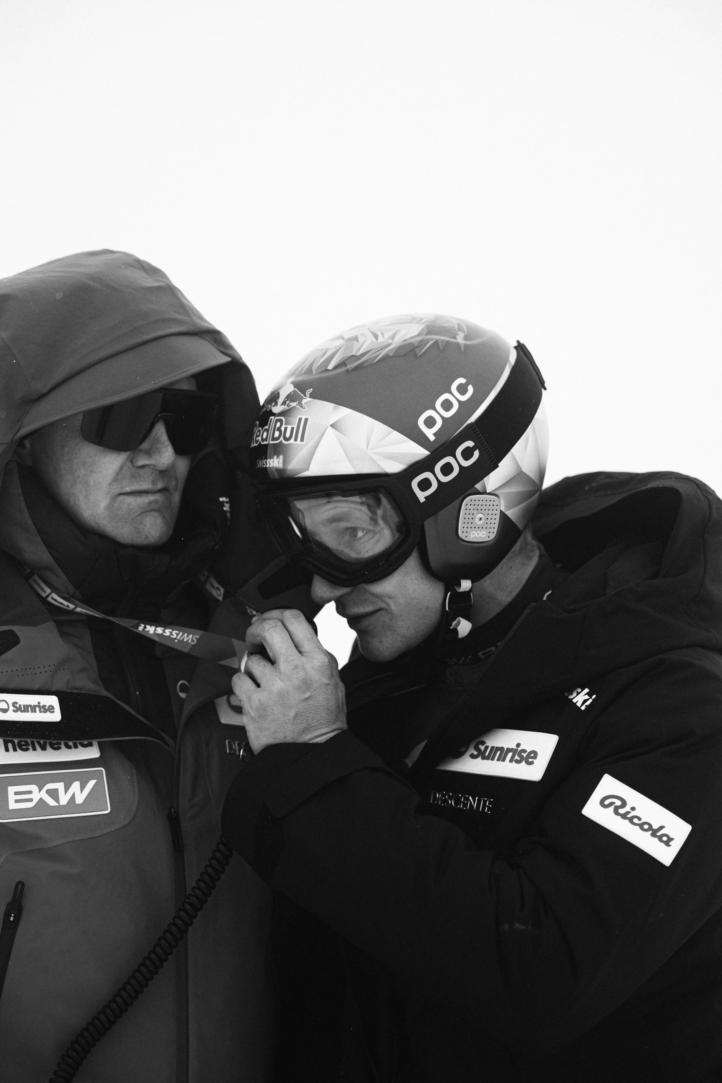 Two men, one with a helmet and goggles, and the other wearing a jacket with sponsor logos, share a close moment, possibly a handshake or a fist bump.