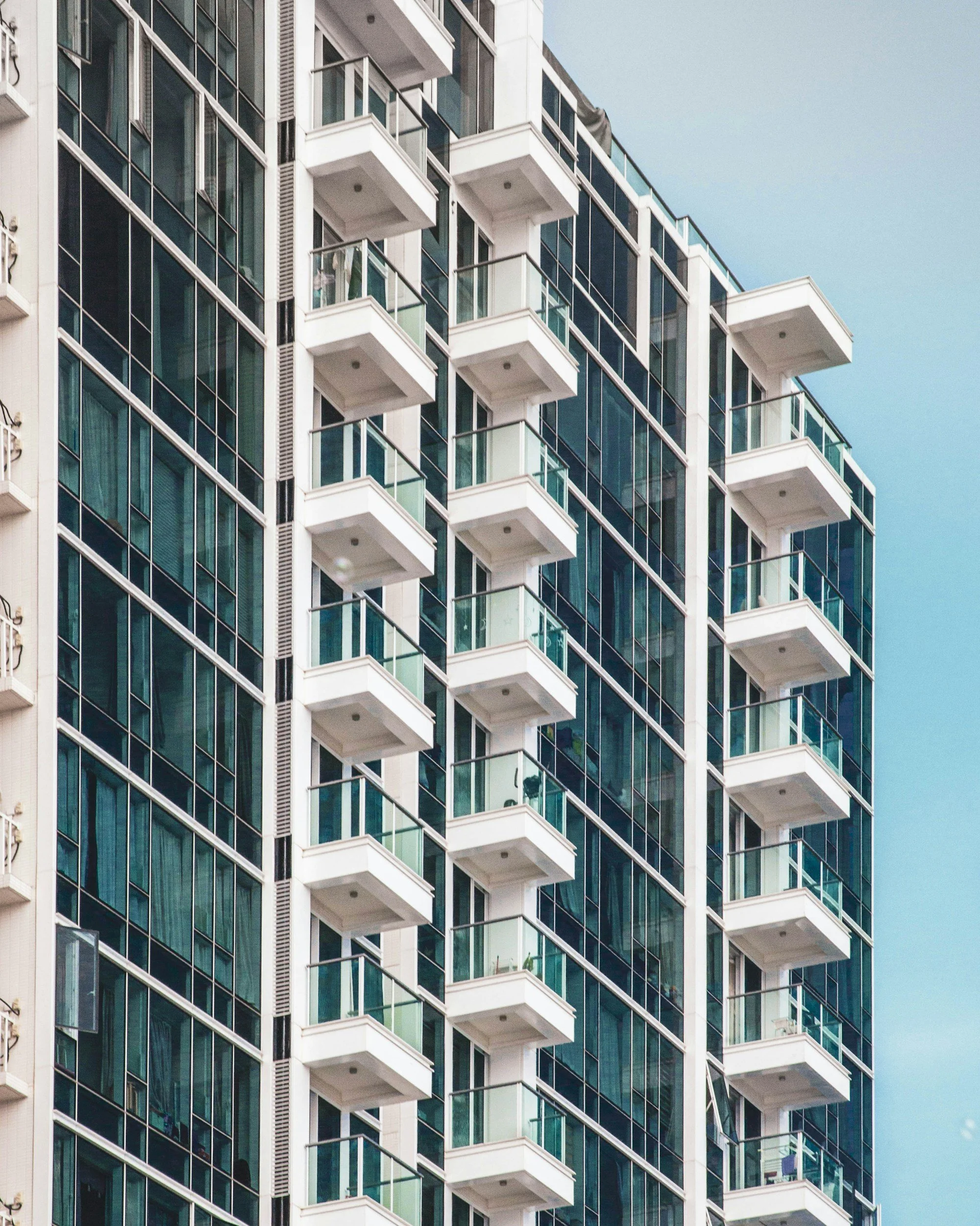 Close-up view of a modern high-rise apartment building with glass windows and balconies, some with glass railings, against a background of a clear blue sky.