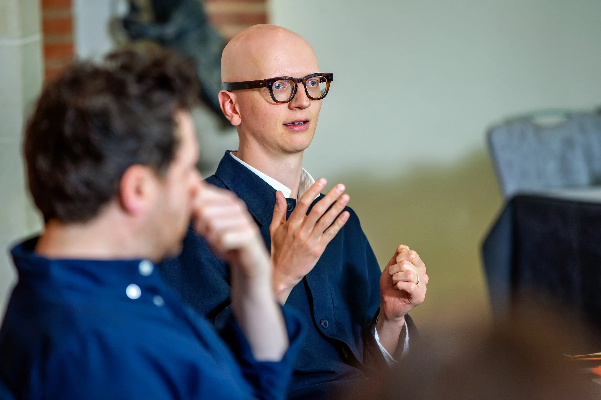 A man with a bald head and glasses is speaking in a meeting, with a woman in front of him listening.