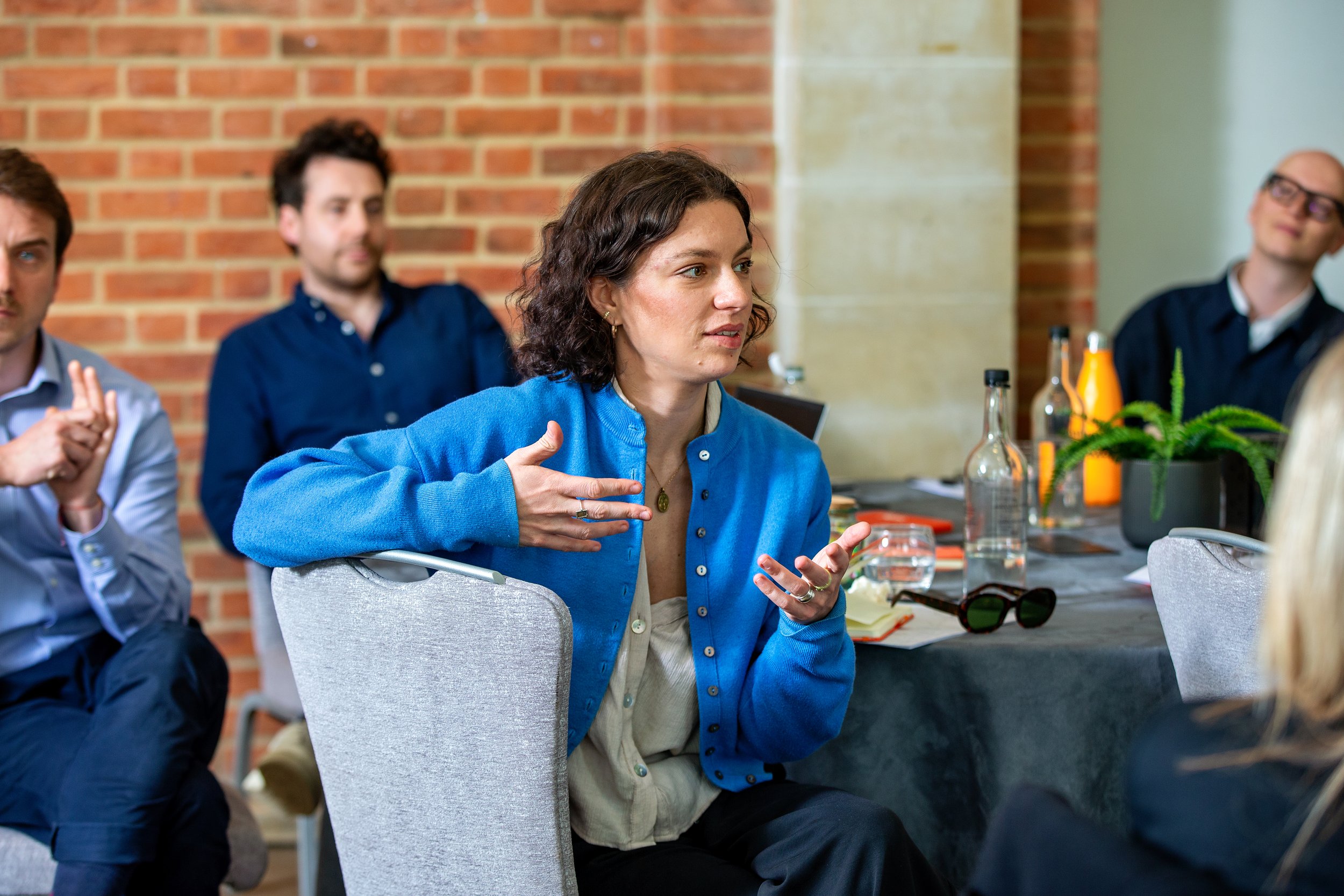 A woman with dark curly hair in a blue jacket speaking and gesturing during a meeting with colleagues in a room with exposed brick wall.