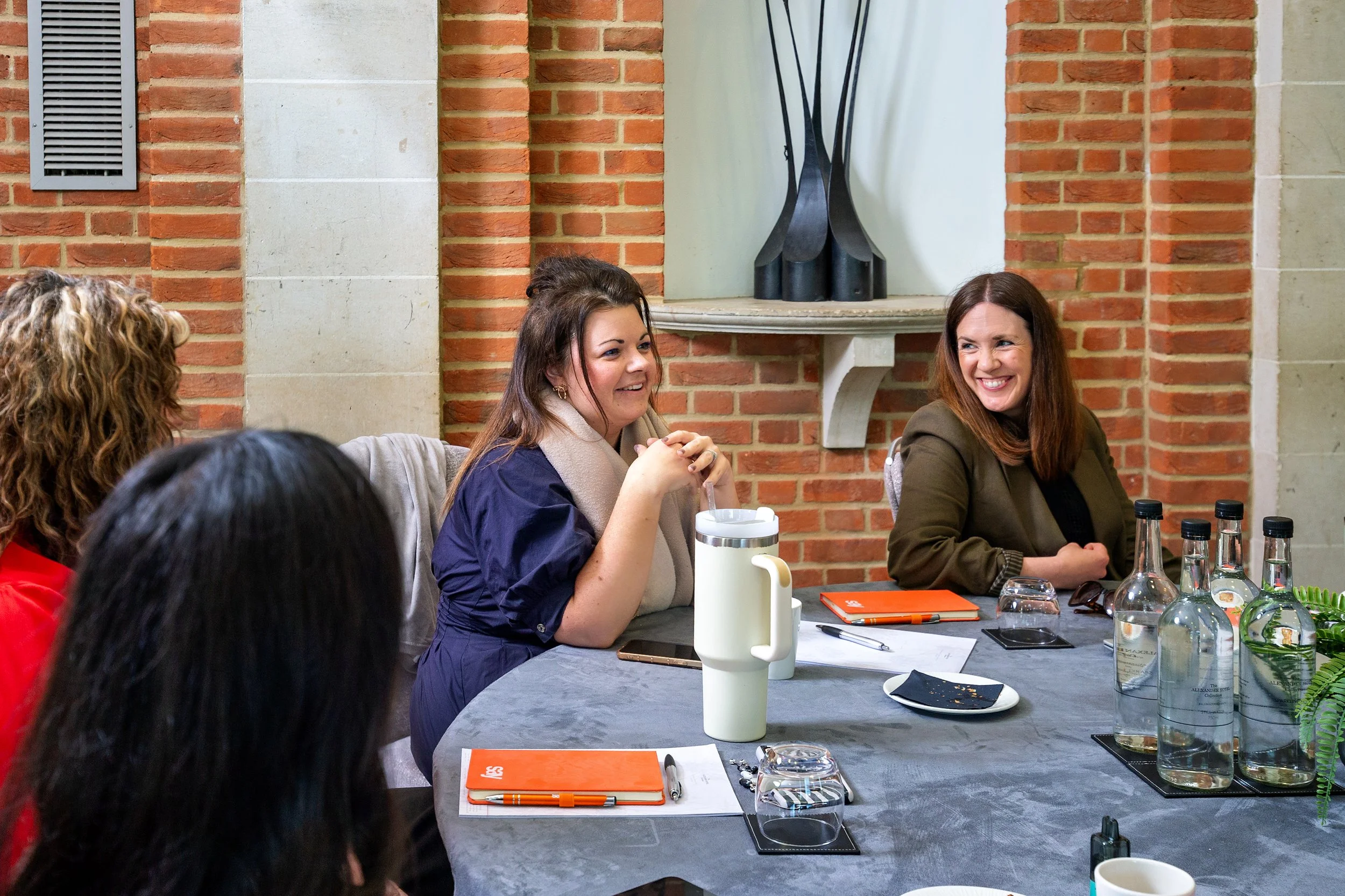 Women sitting at a table in a brick-walled room with drinks, notebooks, and pens, engaged in conversation.