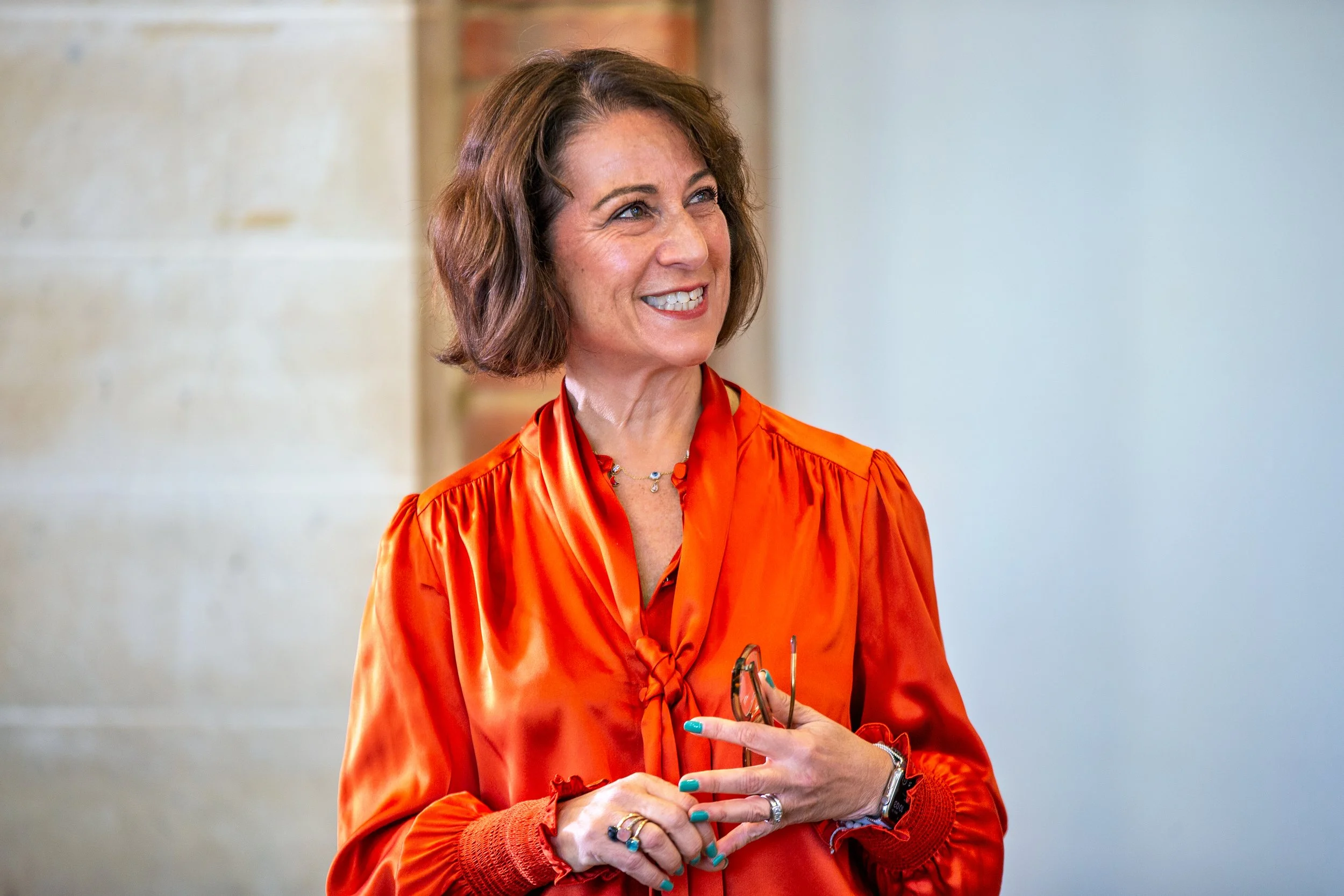 A woman with shoulder-length brown hair wearing an orange satin blouse, holding a pair of eyeglasses, smiling in an indoor setting with a plain wall background.