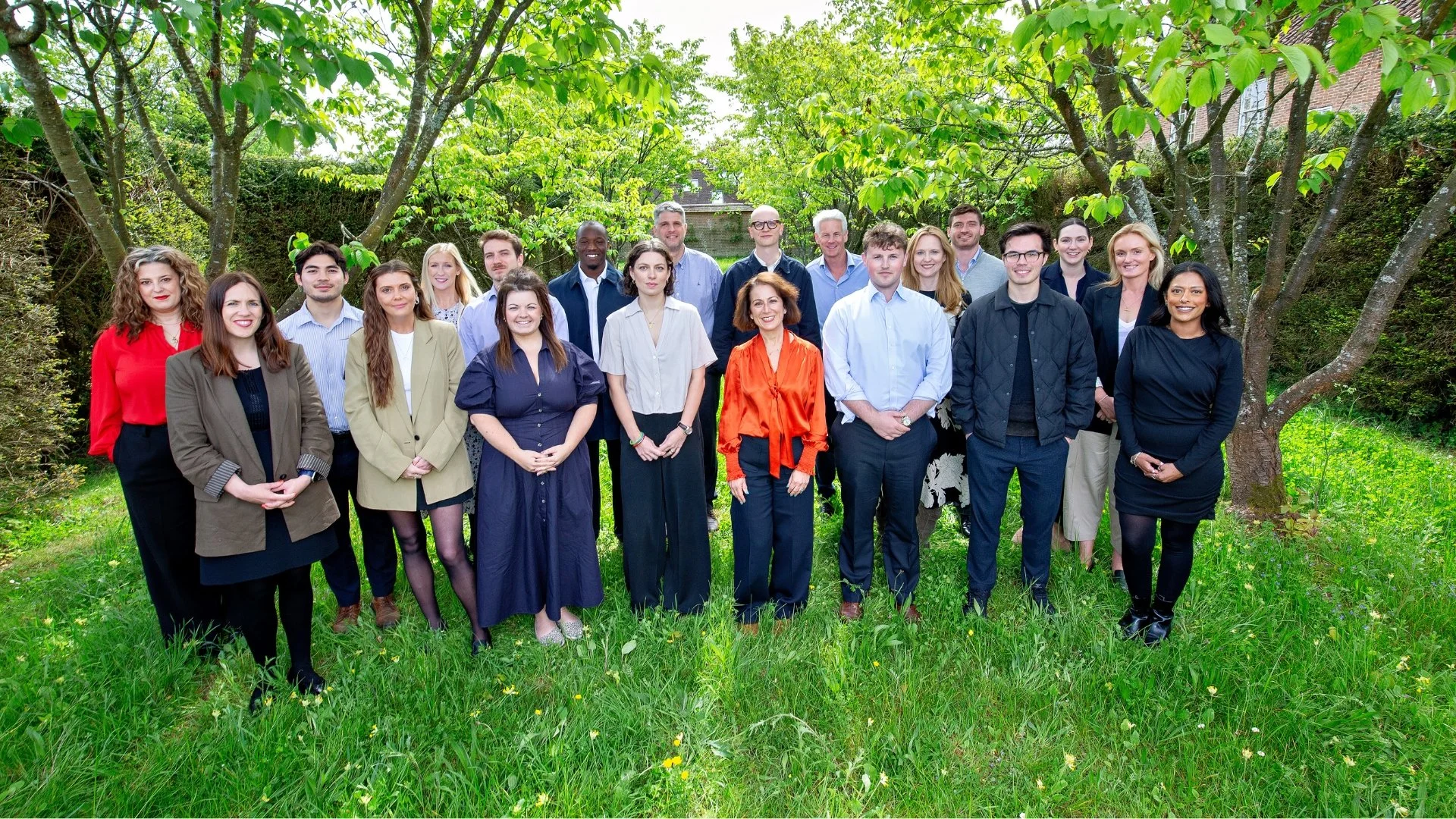 Group of diverse professionals outdoors in a park or garden, standing on grass with trees and a building in the background, smiling at the camera.