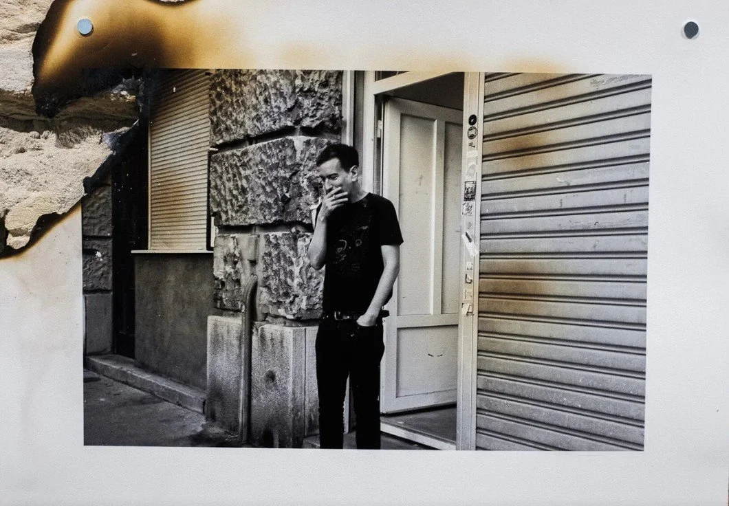Black and white image of a person standing outside a building, smoking a cigarette, near a doorway with stickers.