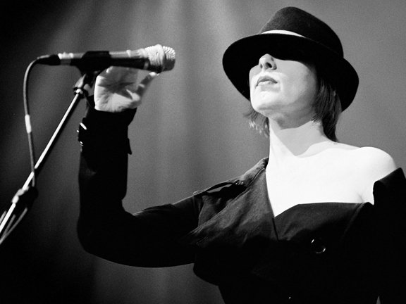 Black-and-white photo of a person wearing a hat, singing into a microphone on stage.
