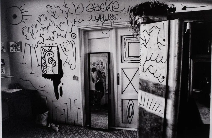Black and white photo of a man sitting in a wicker chair in an art studio with drawings on the wall.