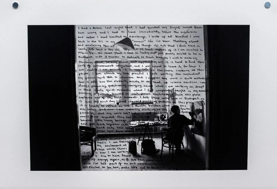 Black and white image of an artist painting in a sparse room with two windows, a hanging lamp, and a table with art supplies.