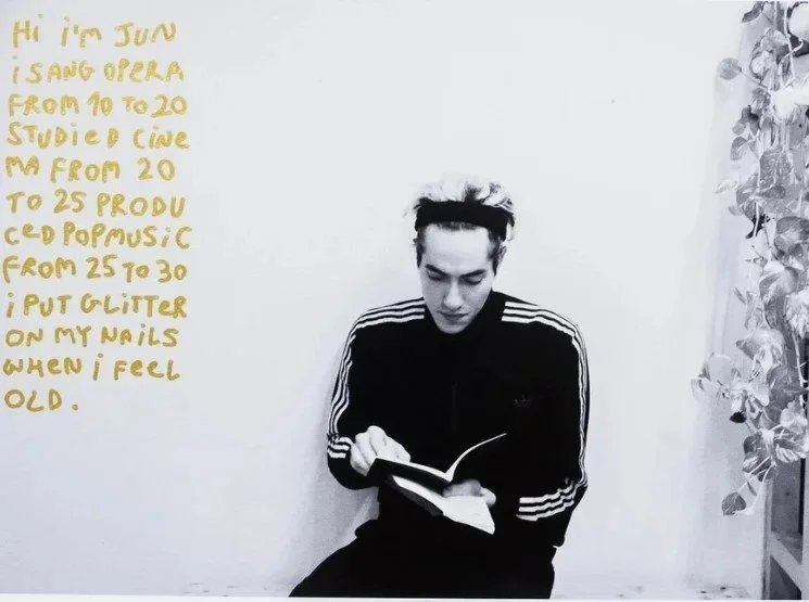 Person reading a book, wearing a striped athletic jacket and headband, sitting on a bench next to a leafy plant in black and white.