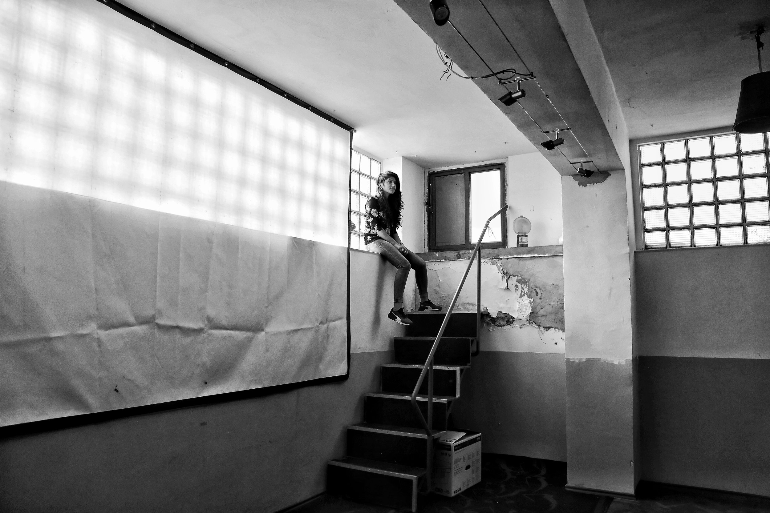 Person sitting on a ledge in a minimalist, industrial-style room with large windows and overhead lights.
