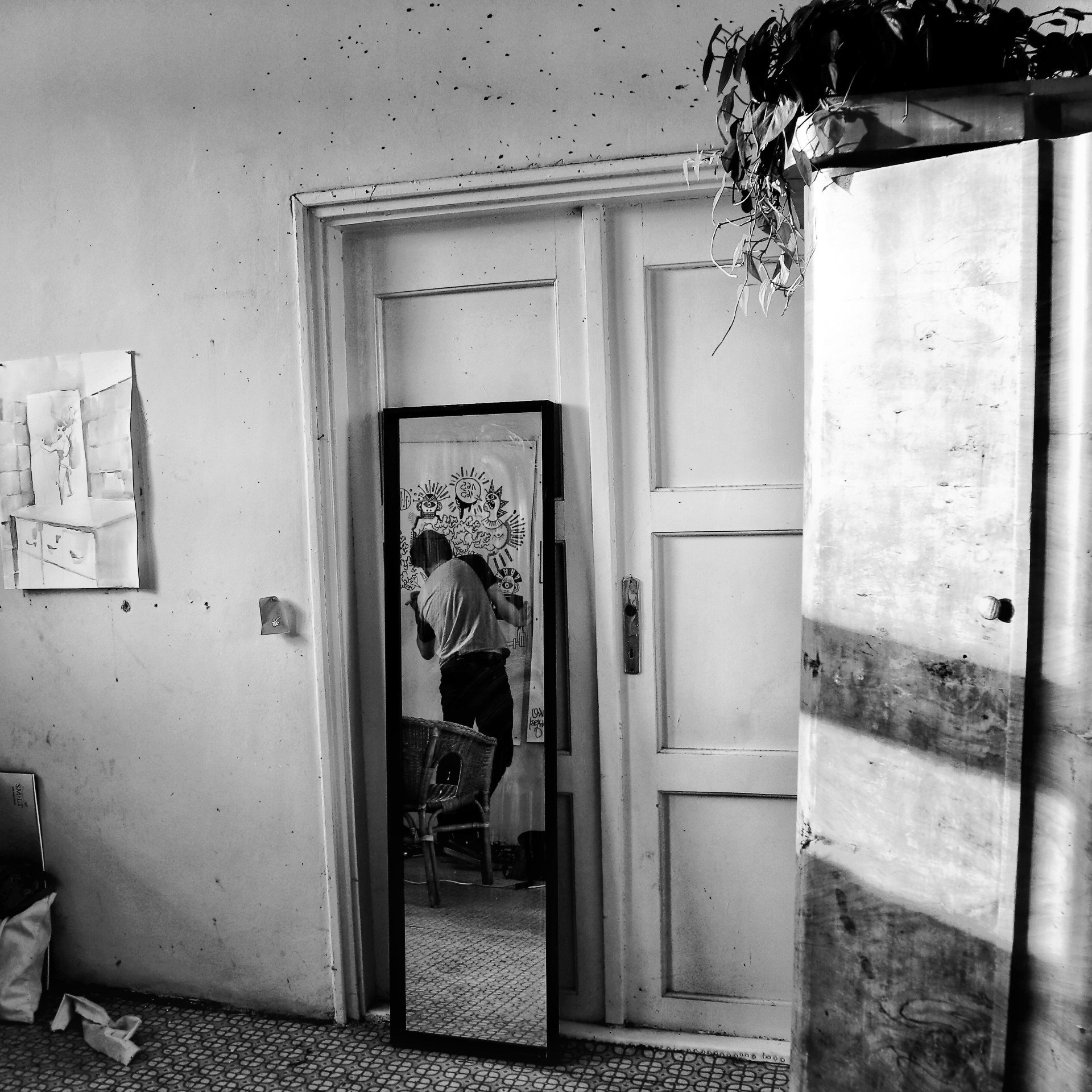 Black and white photo of a man sitting in a wicker chair in an art studio with drawings on the wall.