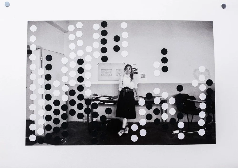 A woman seated beside a desk cluttered with papers and documents, with architectural drawings taped to the wall.