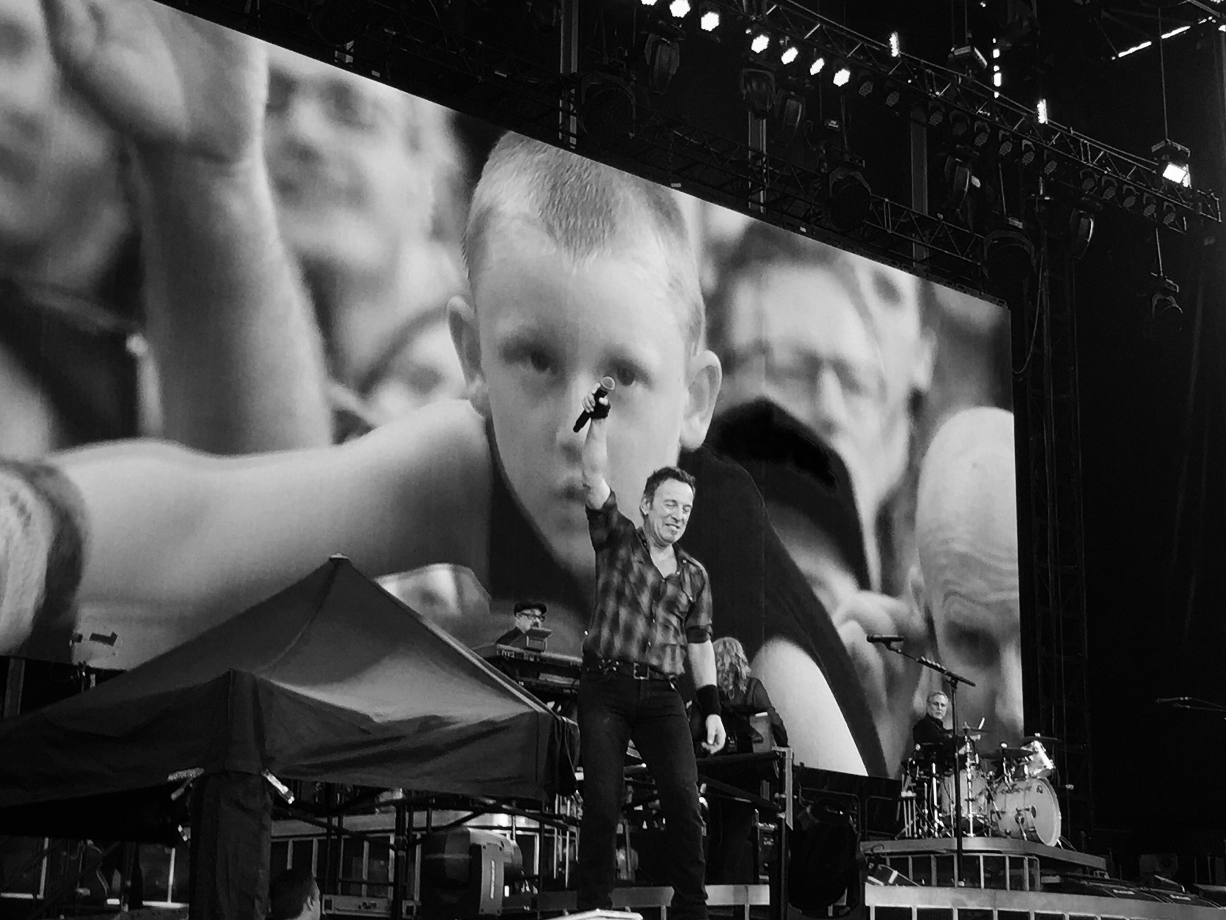 Black and white photo of a musician on stage holding a microphone, with a large screen displaying an audience behind. A band setup including a drummer is visible.