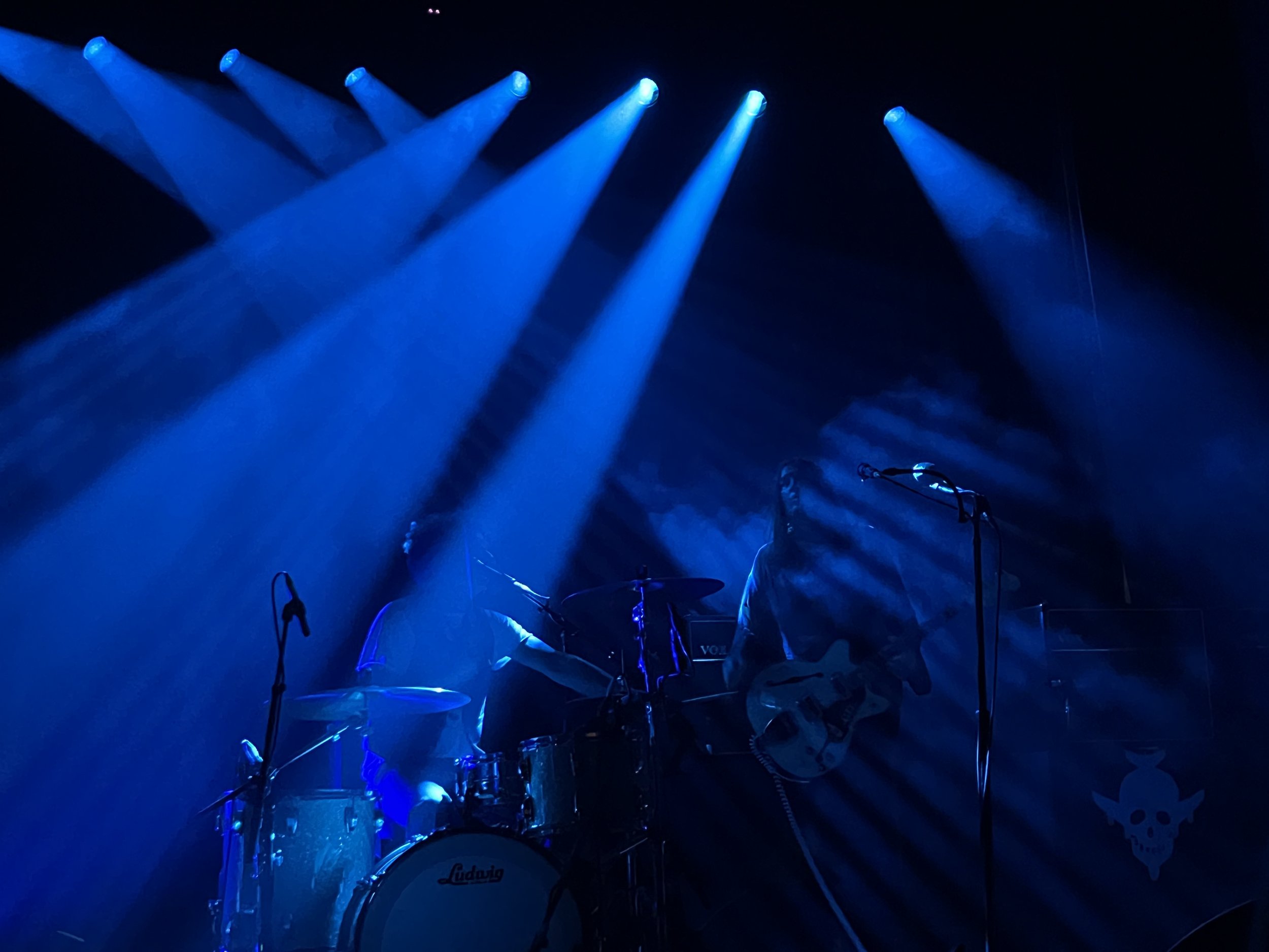 A dimly lit stage with blue lights, featuring a drummer and a guitarist playing under spotlights.