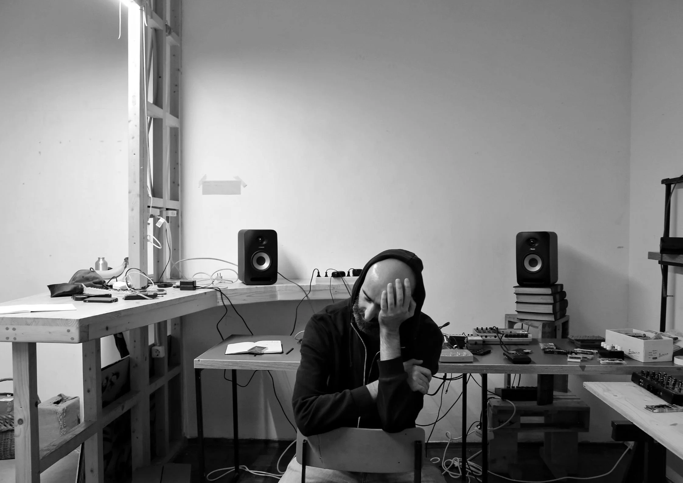 Person in a hoodie working in a cluttered workshop, adjusting audio equipment connected to speakers on a desk.