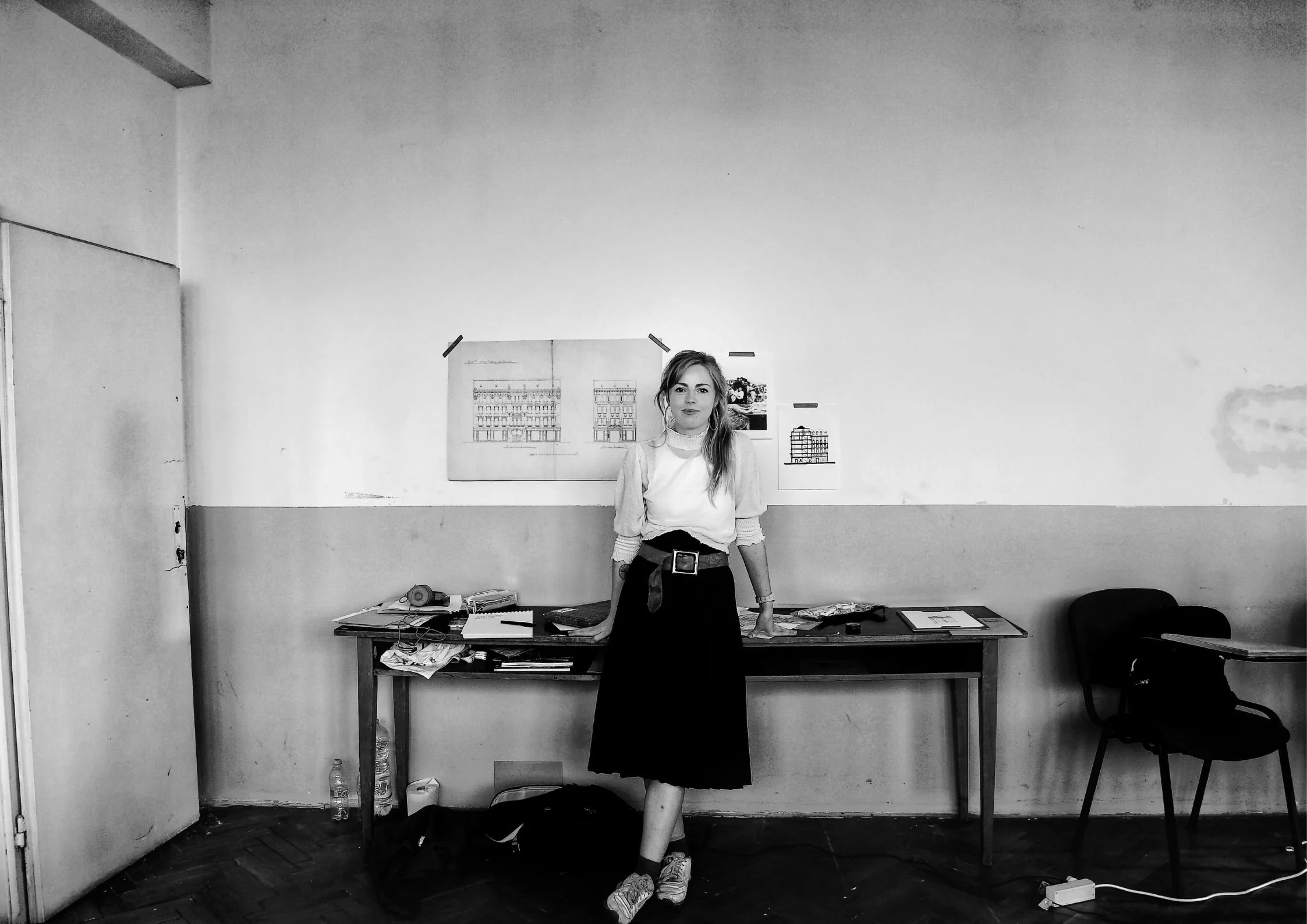 A woman seated beside a desk cluttered with papers and documents, with architectural drawings taped to the wall.