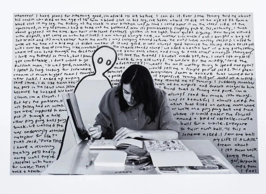 Black and white photo of a person sitting at a table, drawing in a sketchbook with art supplies scattered around.