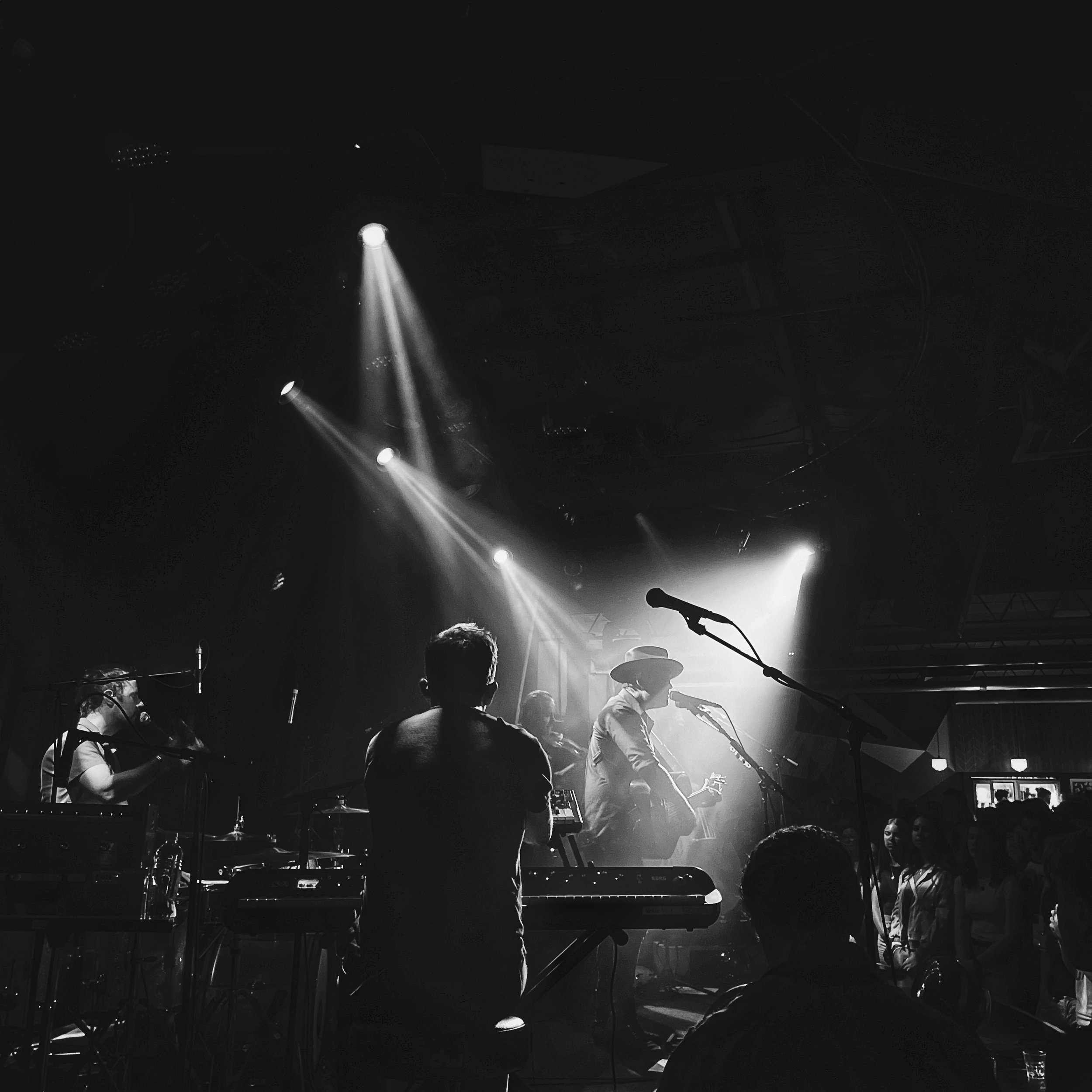 Black and white photo of a live band performing on stage, with musicians playing instruments under spotlights, creating a moody atmosphere.