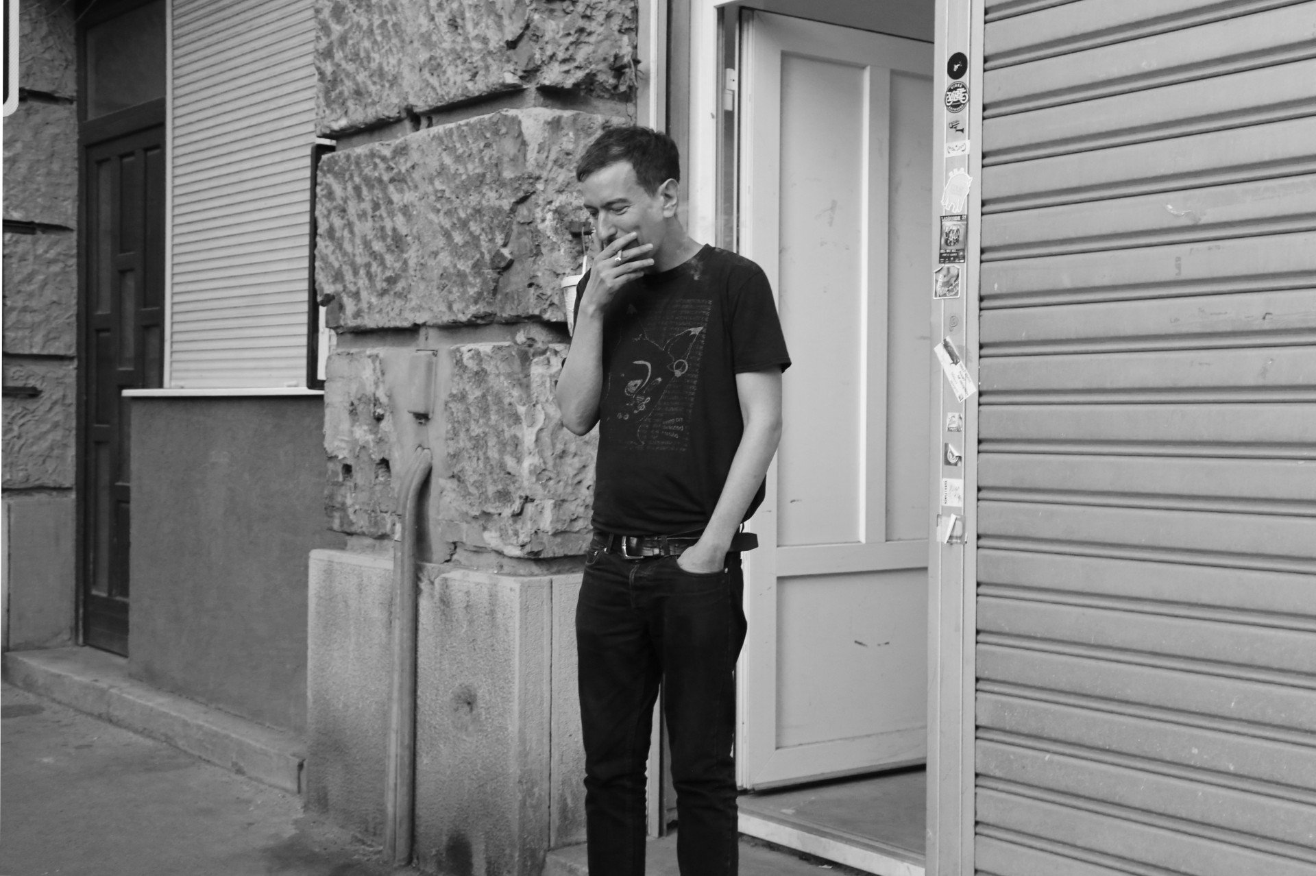 Black and white image of a person standing outside a building, smoking a cigarette, near a doorway with stickers.