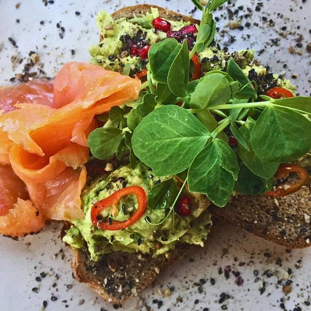 Avocado toast with smoked salmon, pea shoots, red pepper slices, and seasoning on a plate.