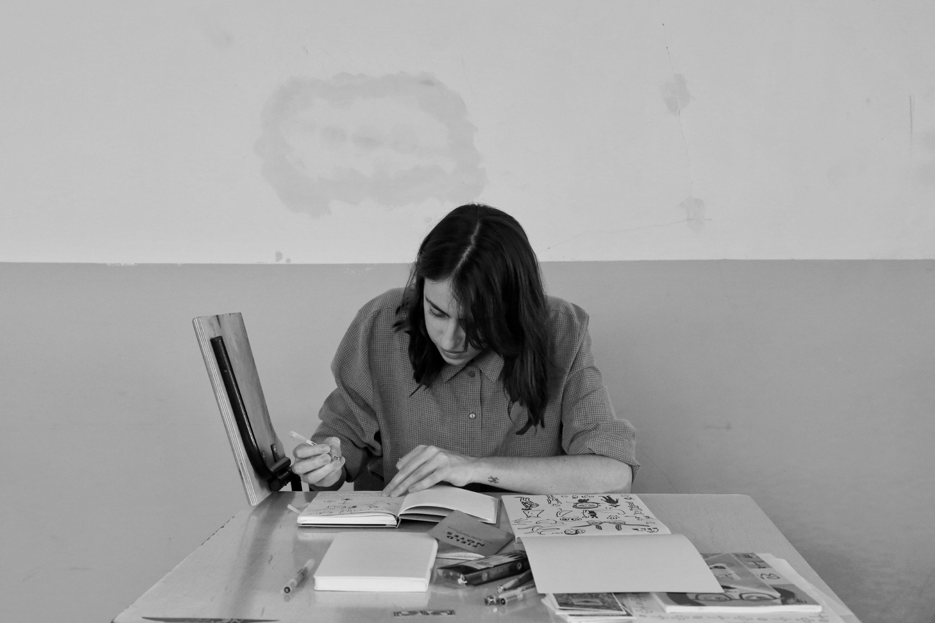 Black and white photo of a person sitting at a table, drawing in a sketchbook with art supplies scattered around.