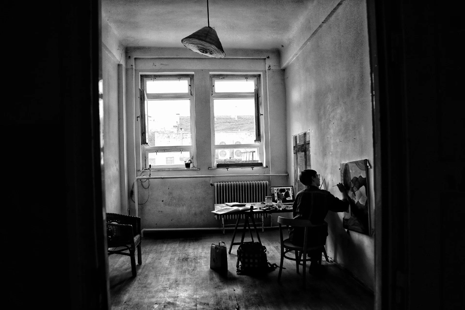 Black and white image of an artist painting in a sparse room with two windows, a hanging lamp, and a table with art supplies.