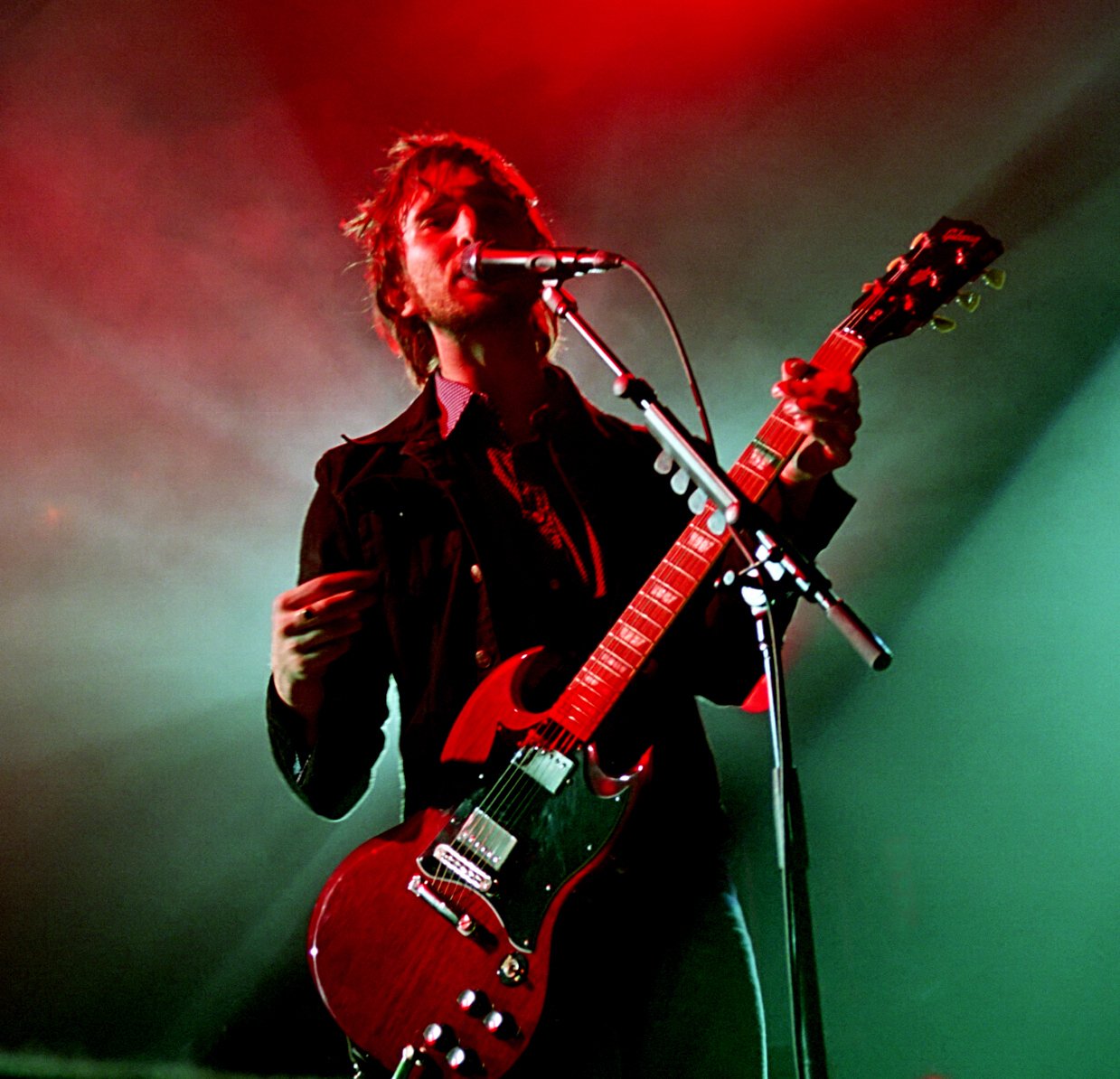 Musician performing on stage with a red electric guitar and microphone, under red and green stage lights.