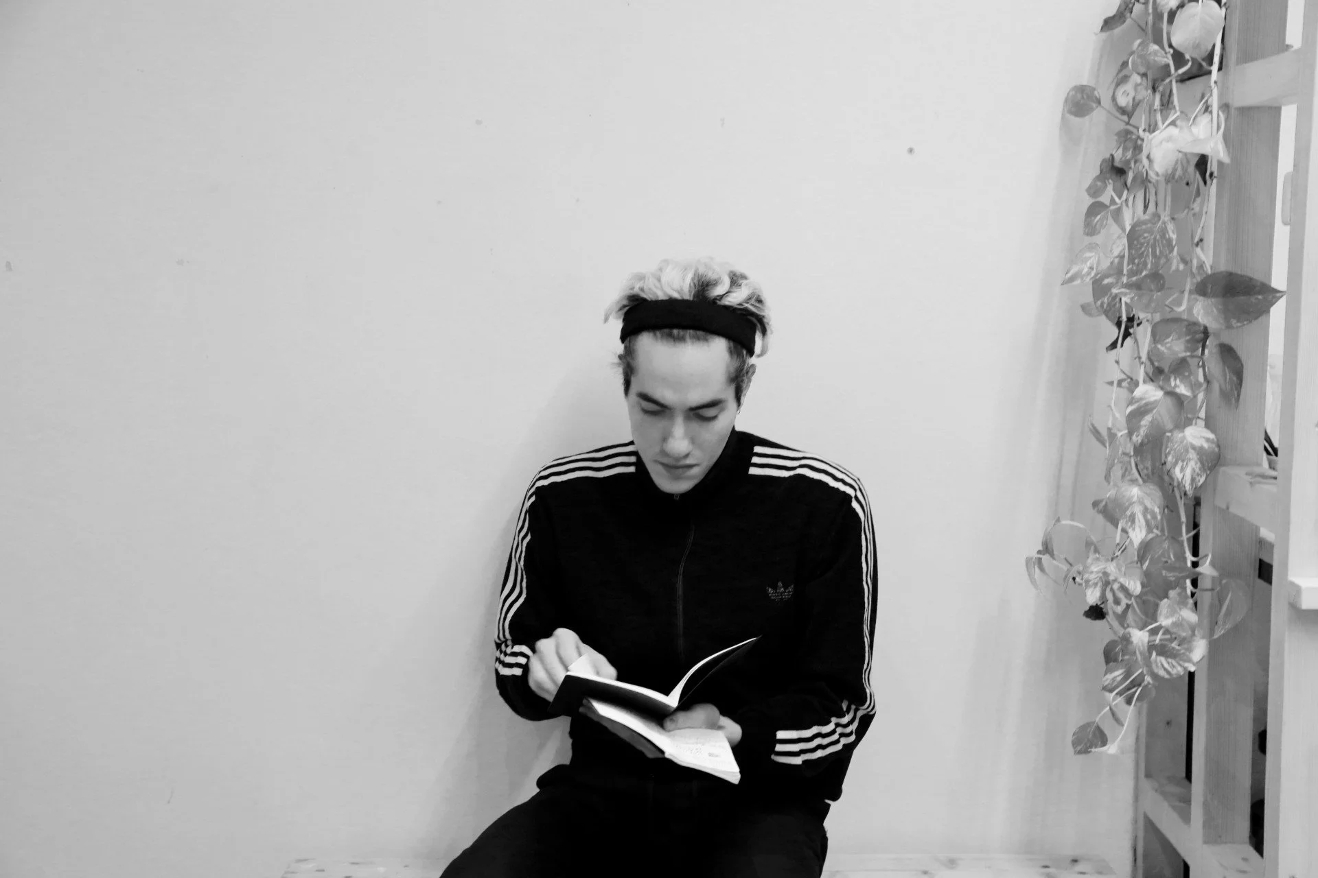 Person reading a book, wearing a striped athletic jacket and headband, sitting on a bench next to a leafy plant in black and white.
