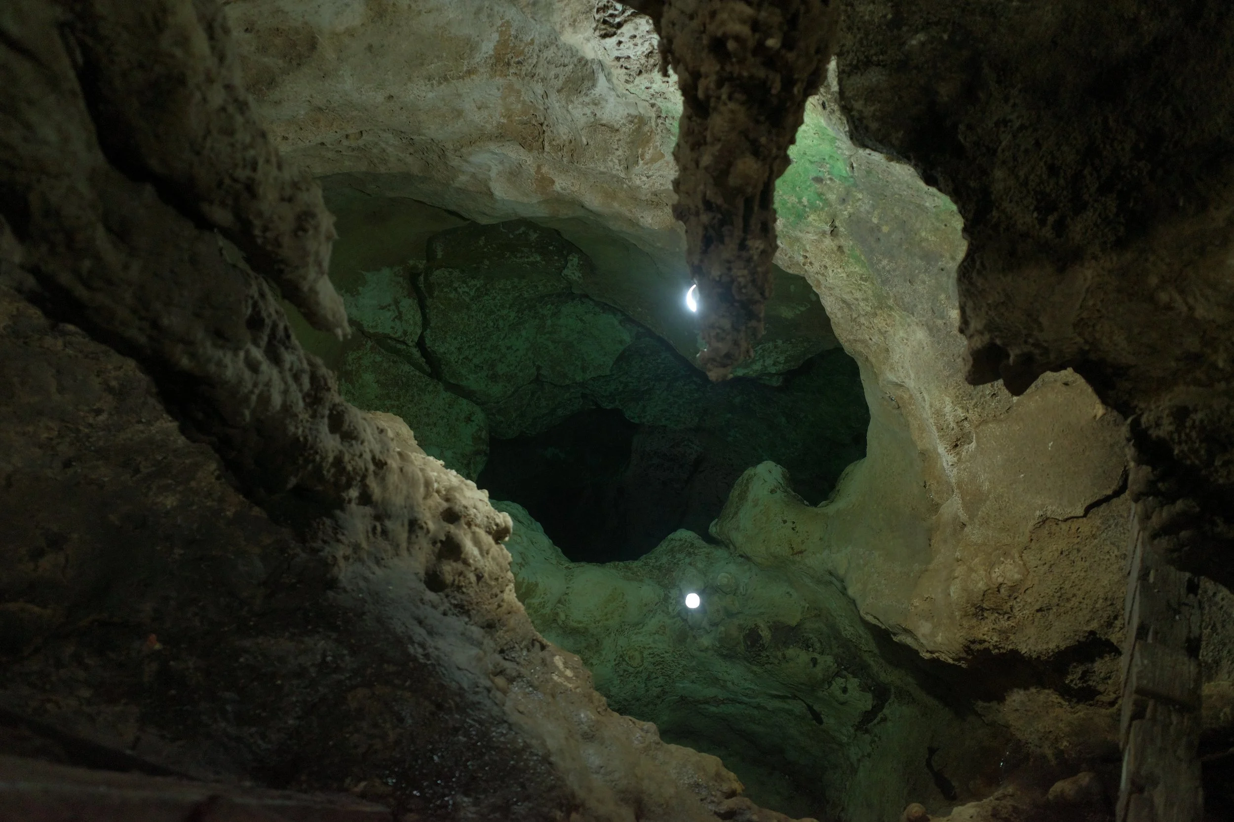 Looking down into a cave through a small opening with sunlight shining through, revealing green moss on the rocks and a dark interior