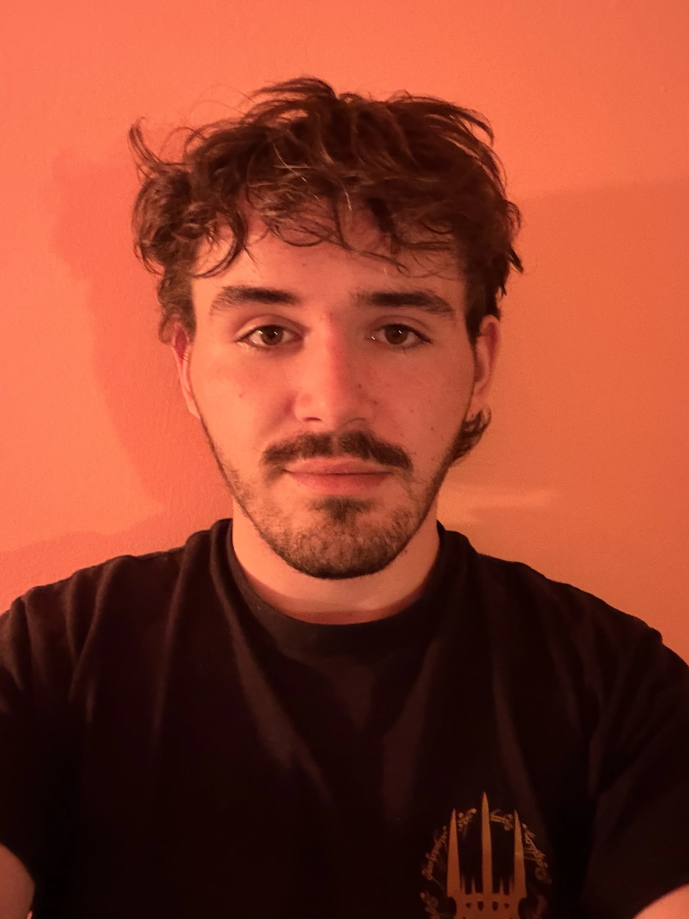 A young man with curly brown hair and a beard, wearing a black T-shirt with a golden graphic, standing against an orange wall.