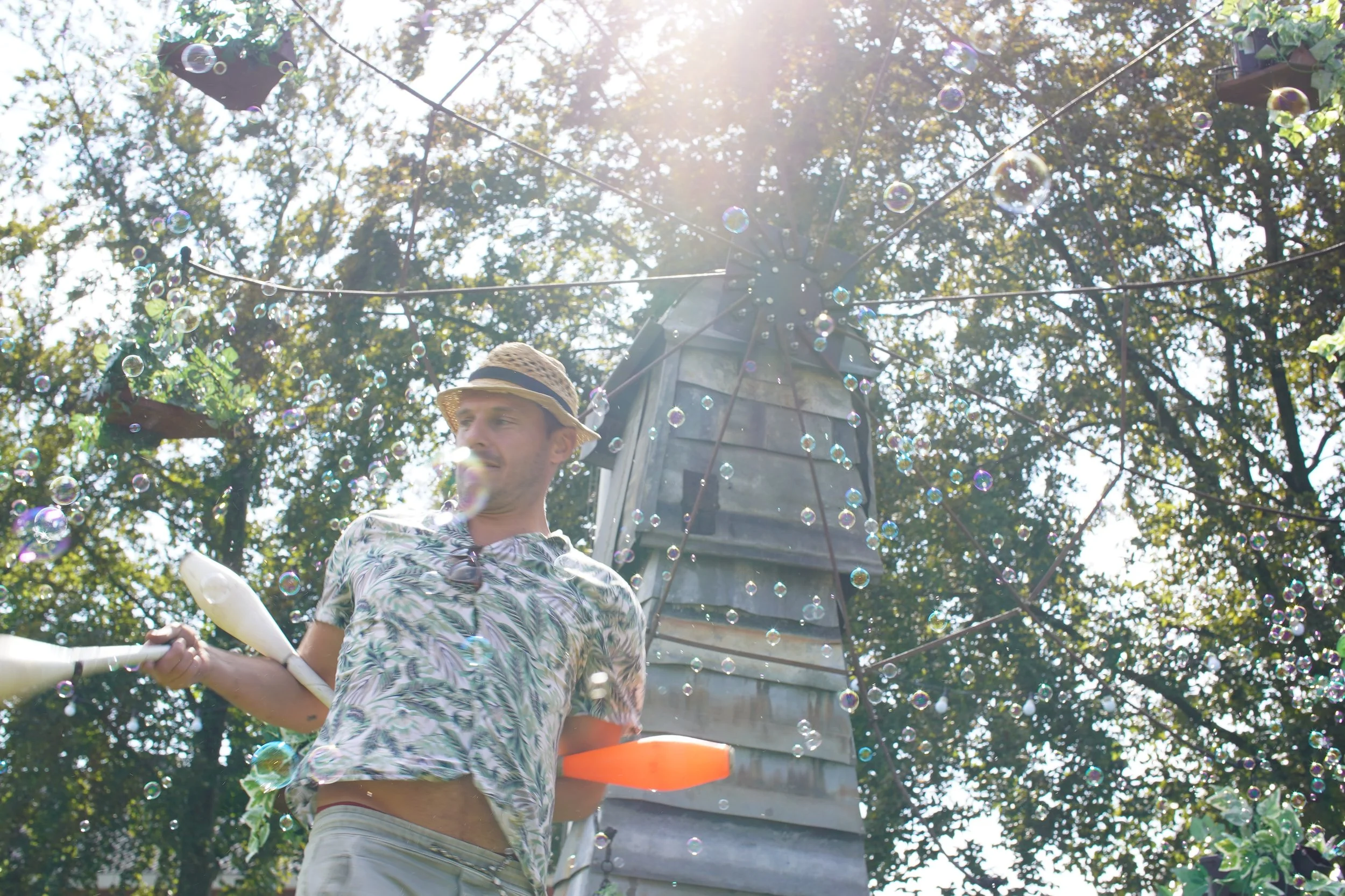 Man met een strooien hoedje die water uit een waterpistool spuit, omringd door zeepbellen, in een bosrijke omgeving met bellenmolen en zonlicht. Bellenmolen, vuurmolen, onthaal, vuuranimatie, vuurspuwer, vuurartiest, vuurshow.