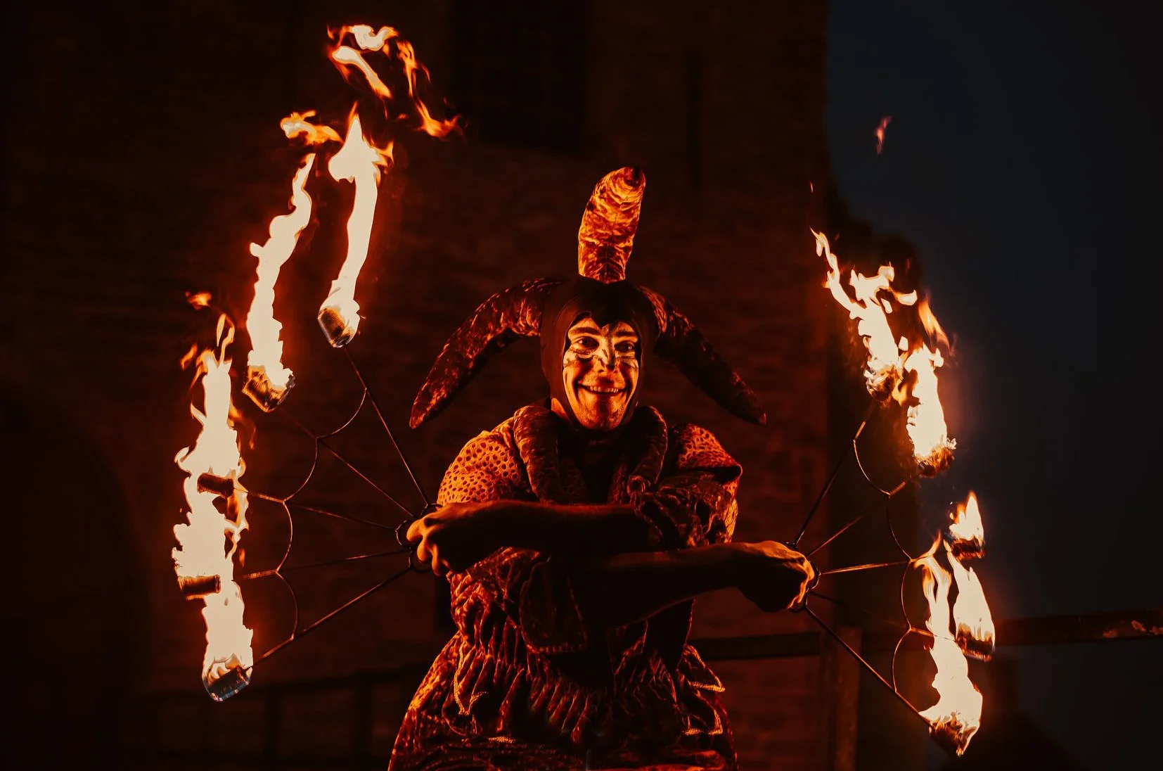 Persoon in een feestkostuum met een hoed met hoorns en vuurvishouders, die vuurspuwt tijdens een optreden 's nachts. Bellenmolen, vuurmolen, onthaal, vuuranimatie, vuurspuwer, vuurartiest, vuurshow.