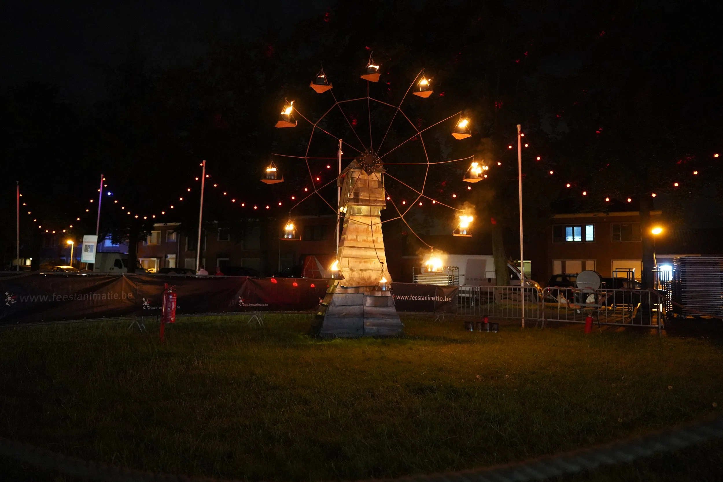 een vuurwiel kunstwerk met brandende lantarens op een grasveld 's nachts, omringd door een hek en lantaarnpalen. Bellenmolen, vuurmolen, onthaal, vuuranimatie, vuurspuwer, vuurartiest, vuurshow.