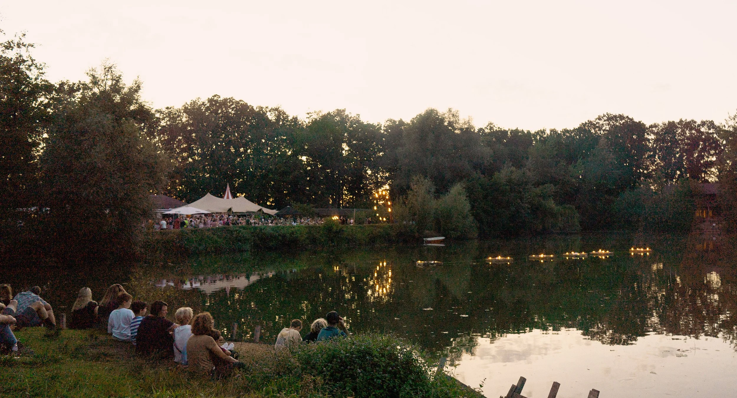 Mensen die langs de oever van een rivier zitten te genieten van de zonsondergang. Aan de overkant is een tent en lichte lichten op het water. Bellenmolen, vuurmolen, onthaal, vuuranimatie, vuurspuwer, vuurartiest, vuurshow.