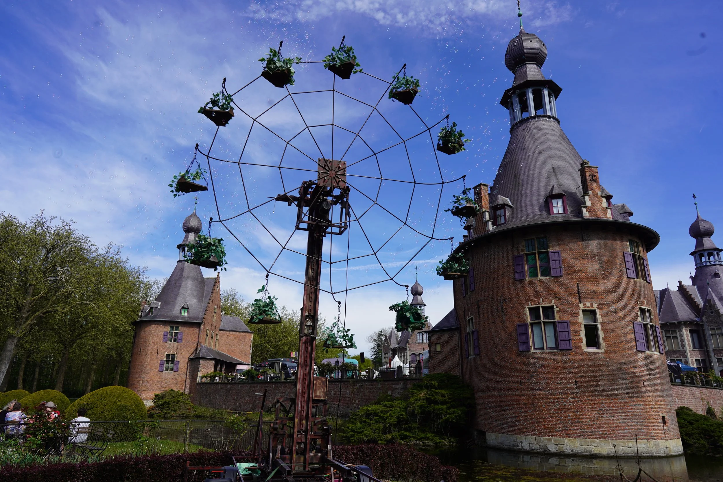 Een oud kasteel met ronde torens en paarse raampwindows, omgeven door groene bomen en een vijver. In de tuin staat een grote ouderwetse draaimolen met een spinnenweb-achtige structuur, waarop bloemenpotten hangen.