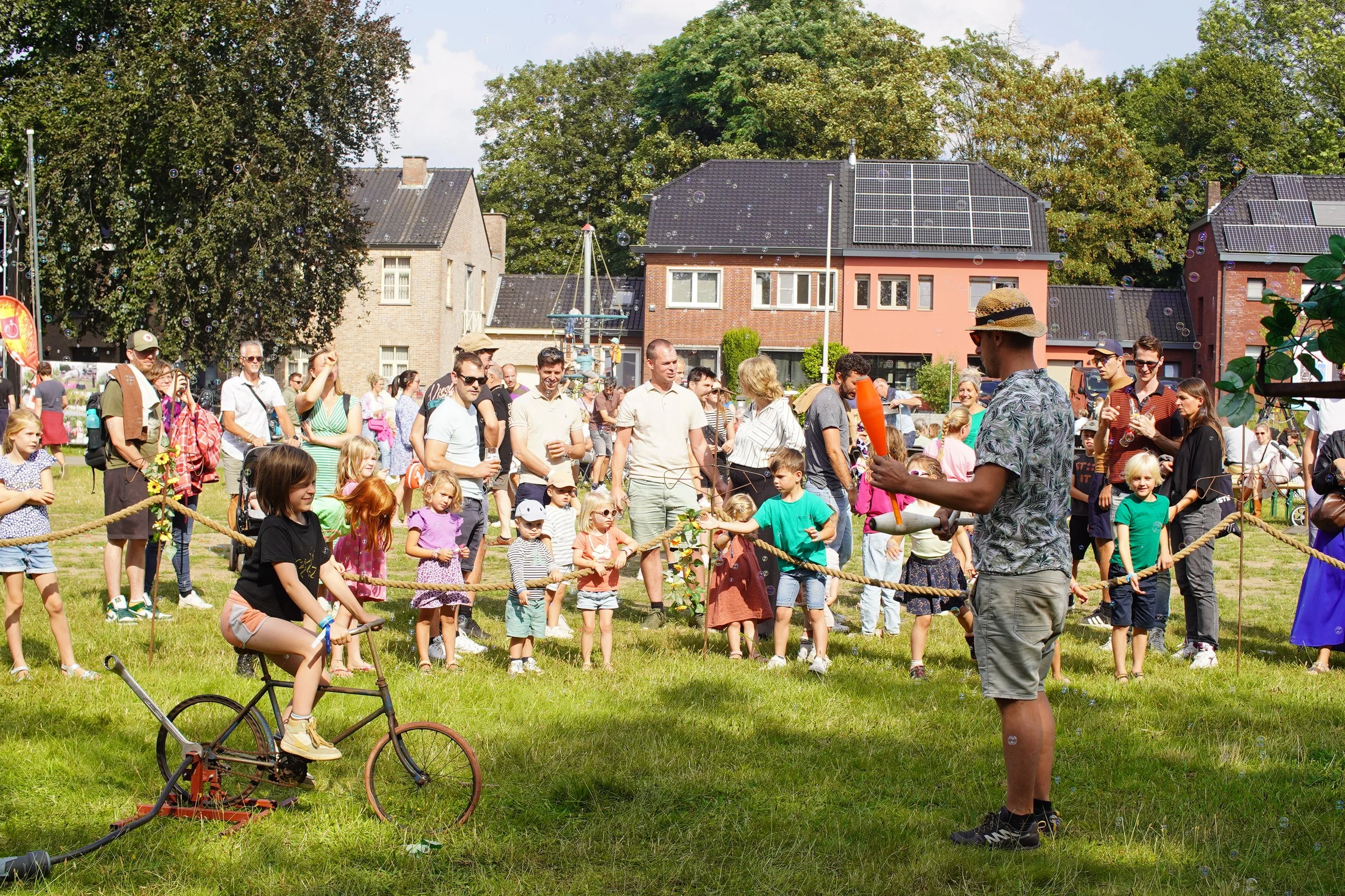 Kinderen en ouders op een grasveld, die naar een clown of entertainer kijken die een ballonfiguur maakt tijdens een festival of evenement op een zonnige dag in de buurt van huizen.