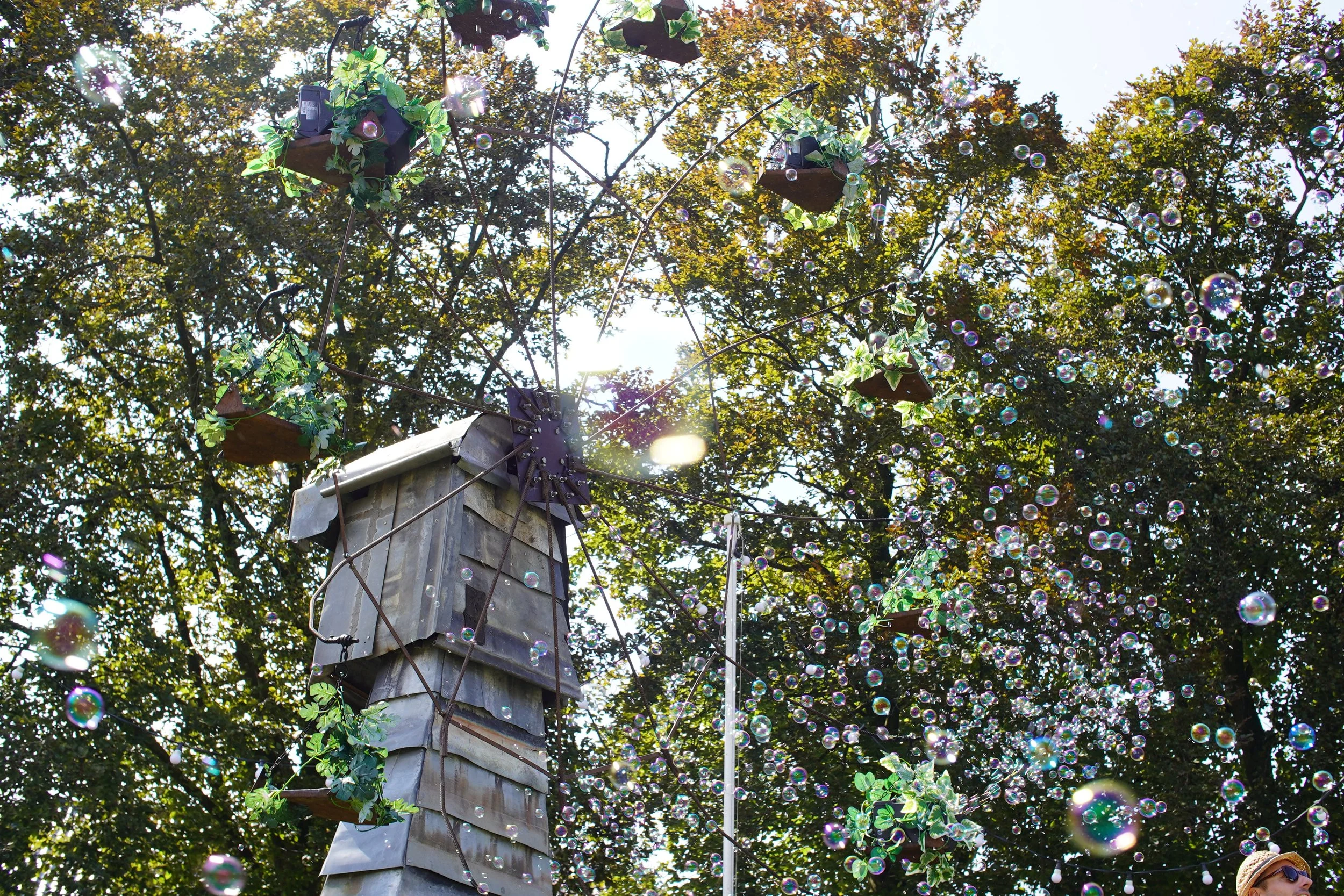 Een windmolen met acht wieken versierd met groene bladeren en planten, onder een heldere lucht met zonnestralen, omgeven door bomen en bellen in de lucht. Bellenmolen, vuurmolen, onthaal, vuuranimatie, vuurspuwer, vuurartiest, vuurshow.