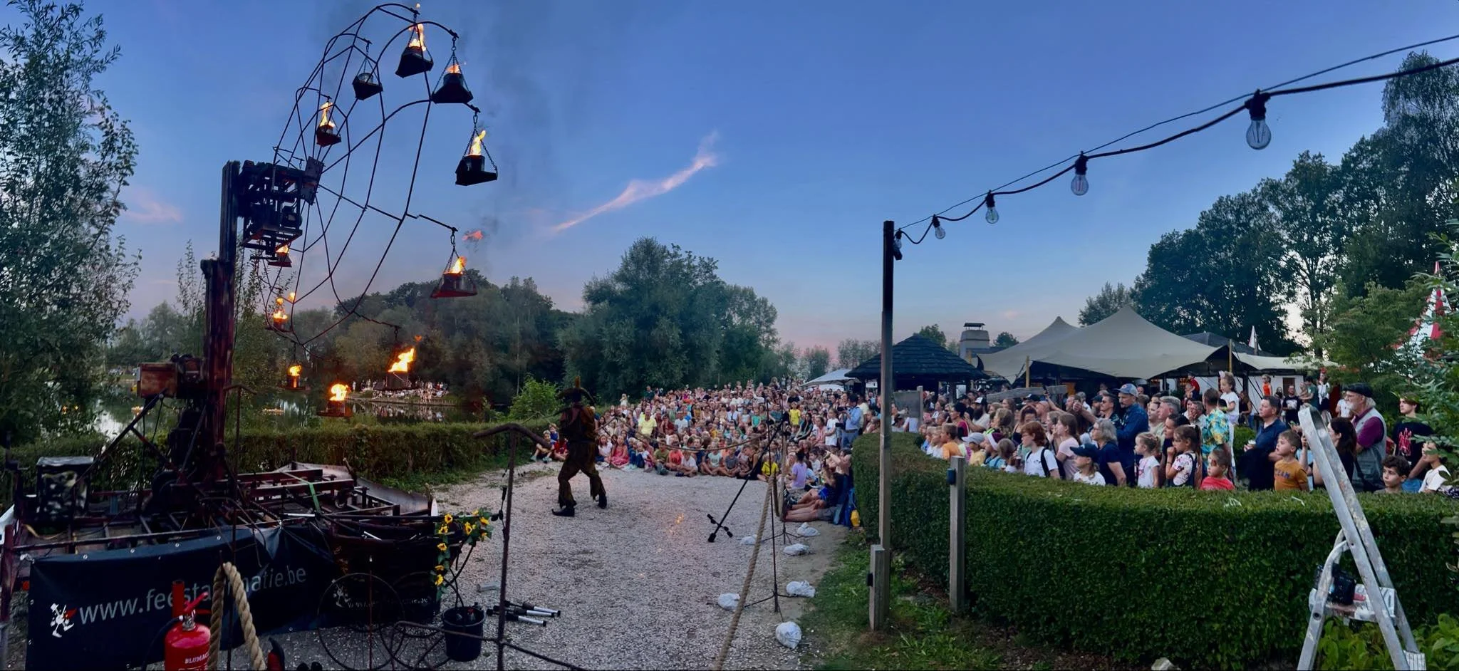 Een openlucht voorstelling met een groot publiek onder een blauwe avondhemel, omgeven door bomen en tents, met een exhibitie van een vuurdier en een kerel in middeleeuwse kleding.
